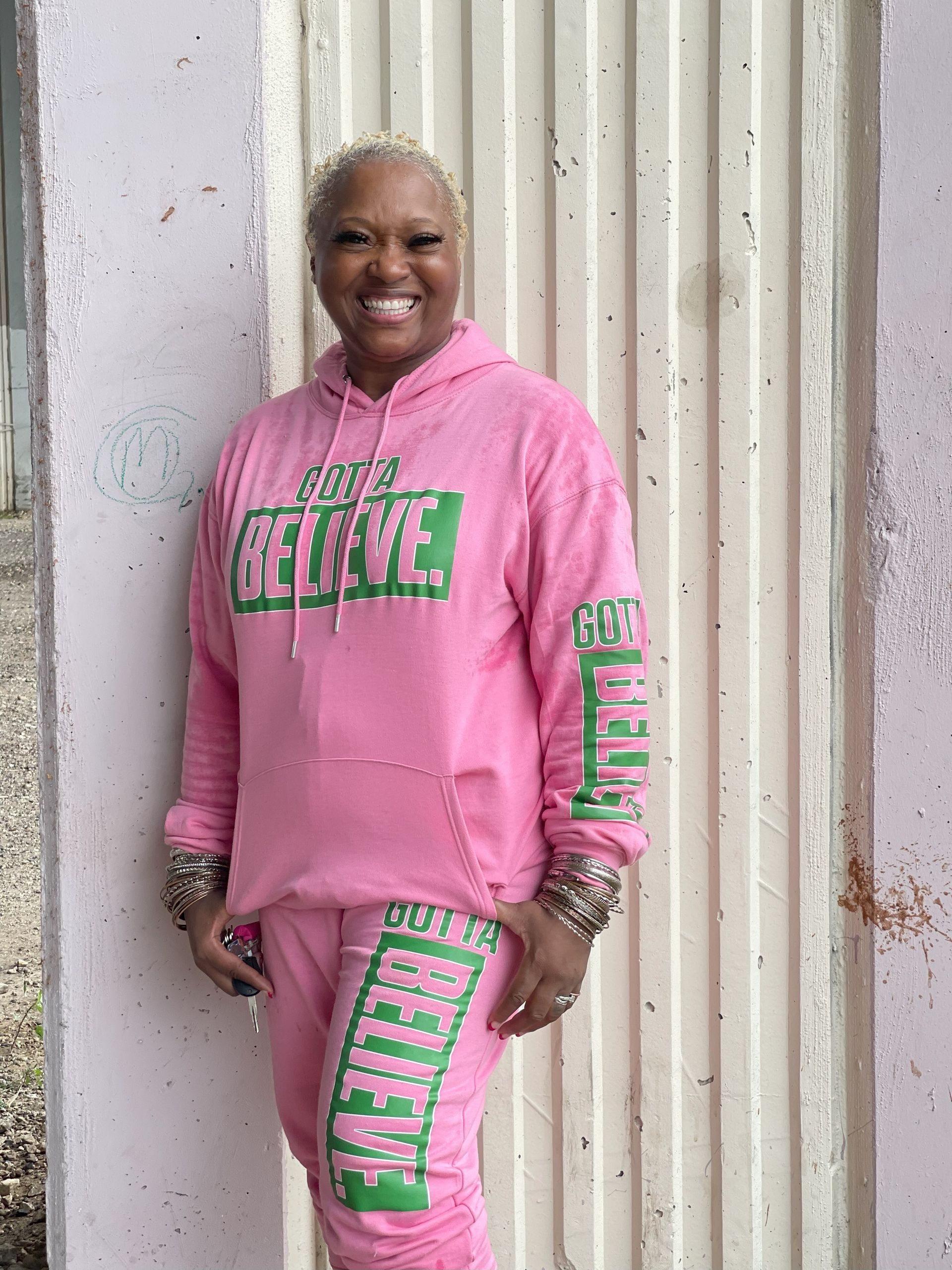 A woman wearing a pink hoodie and pink sweatpants is standing in front of a wall.