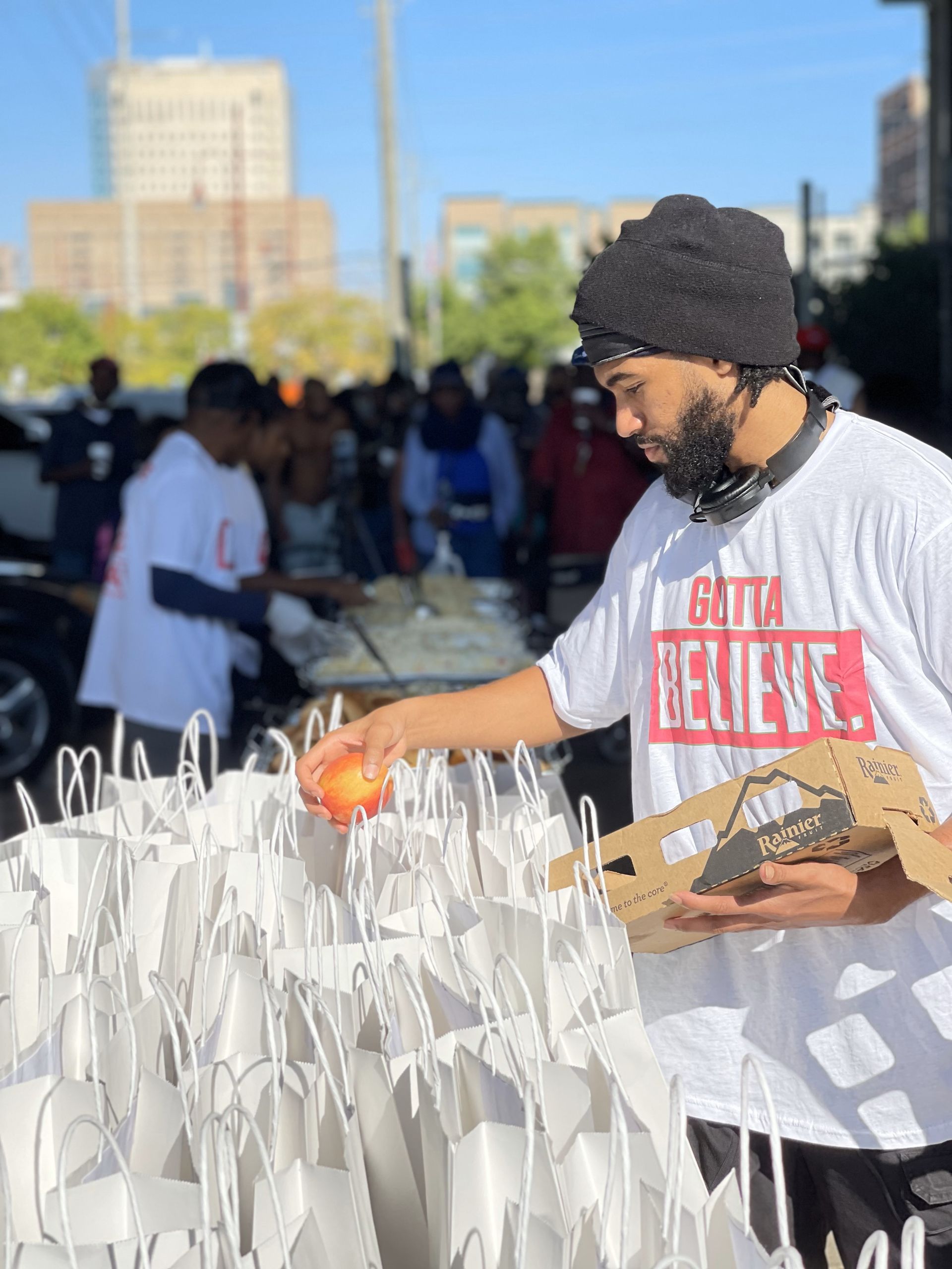 A man in a white shirt with the word believe on it is holding a box of food.