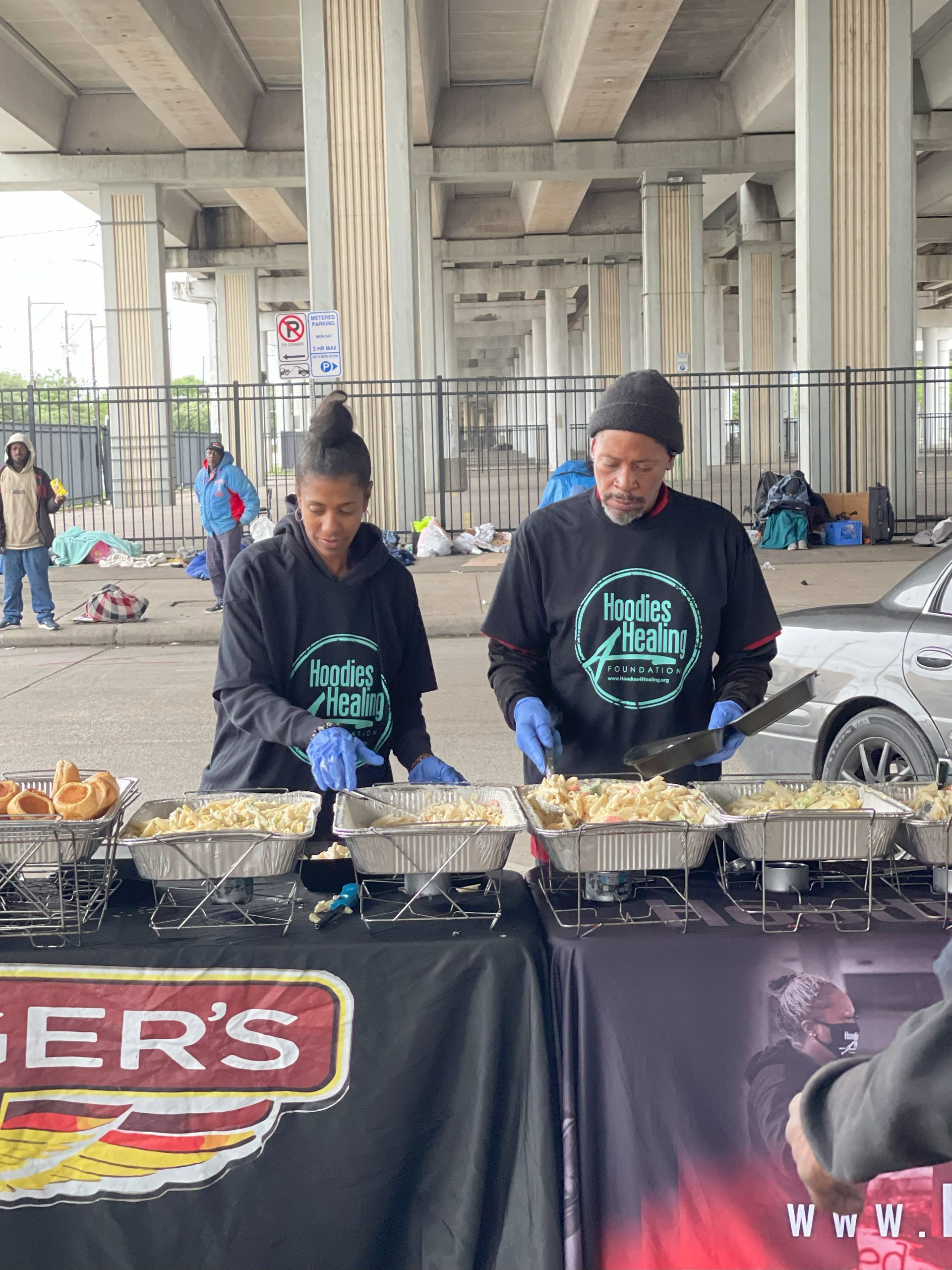 Two people are cooking food at a table under a bridge.