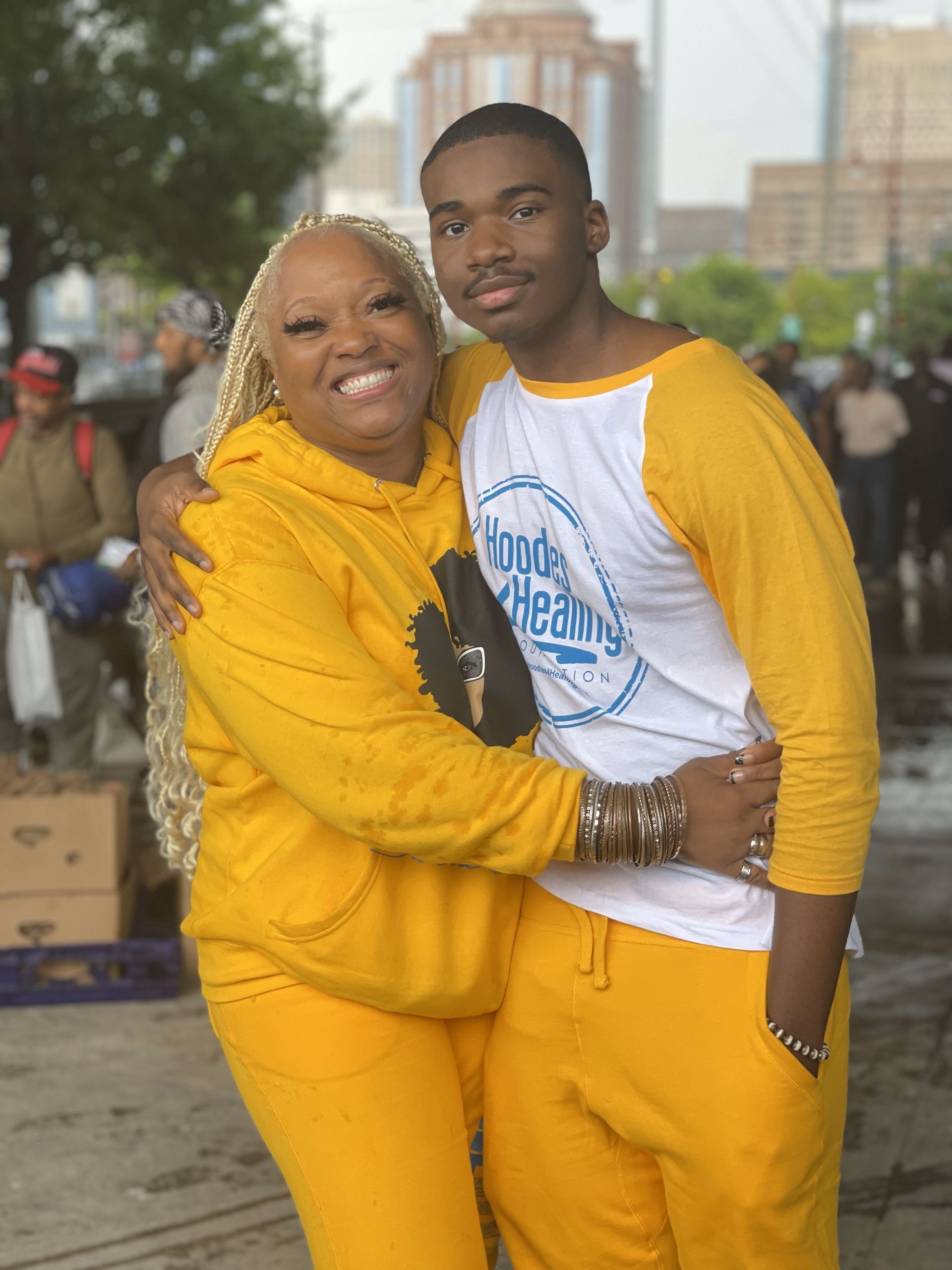 A woman in a yellow hoodie is hugging a young man in a white shirt.