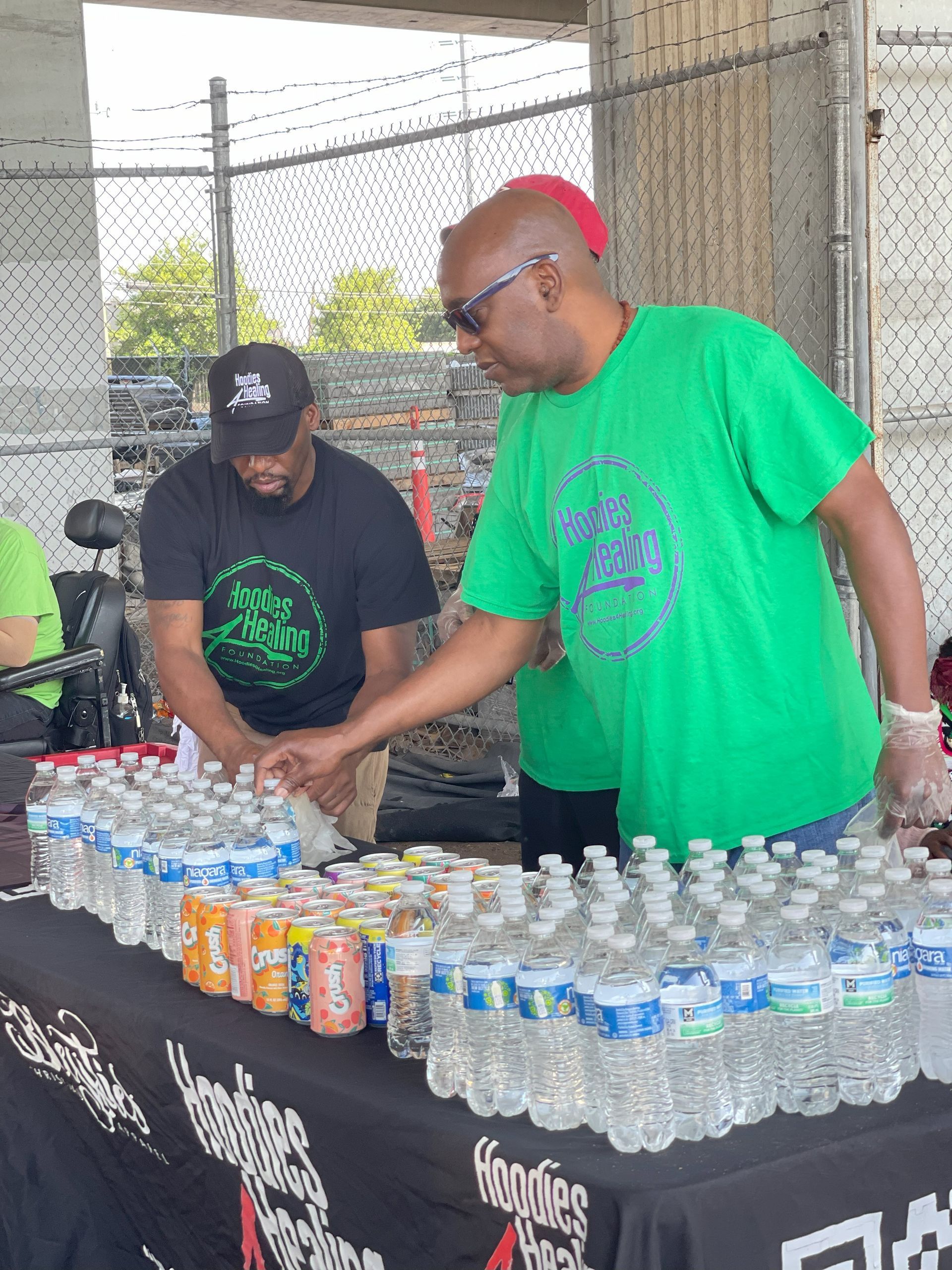A man in a green shirt is standing in front of a table full of water bottles and cans.