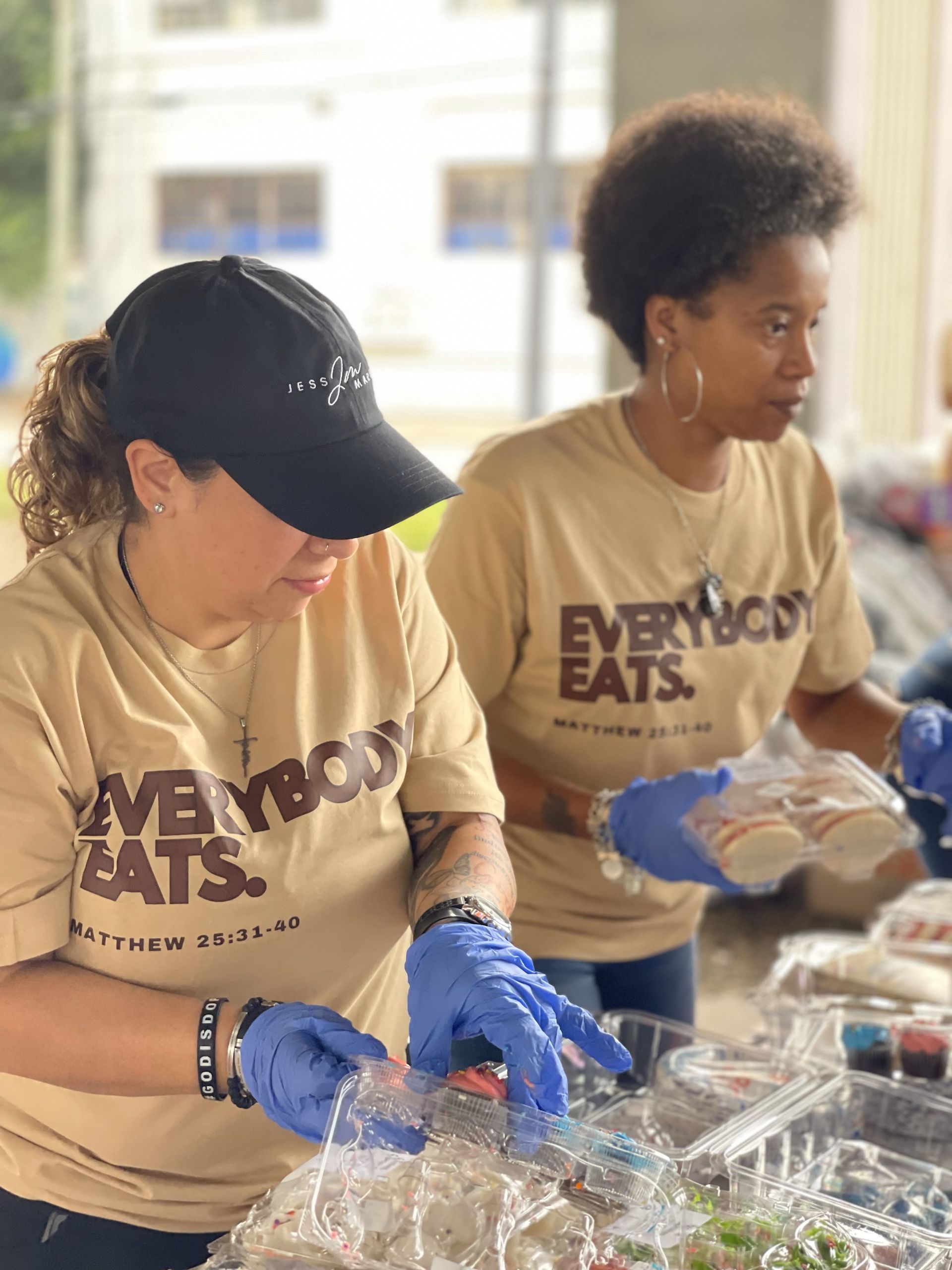 Two women wearing shirts that say everybody eats are preparing food.