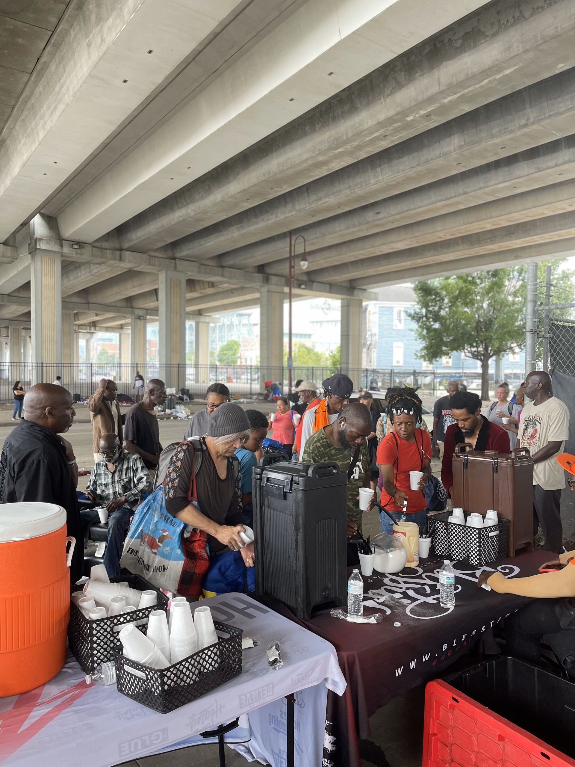 A group of people are standing around tables under a bridge.