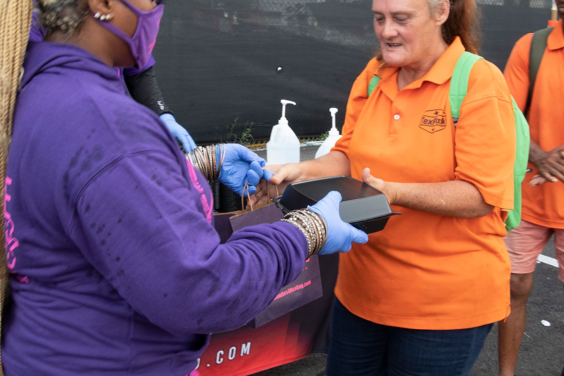 A woman in an orange shirt is giving a box to another woman in a purple shirt.