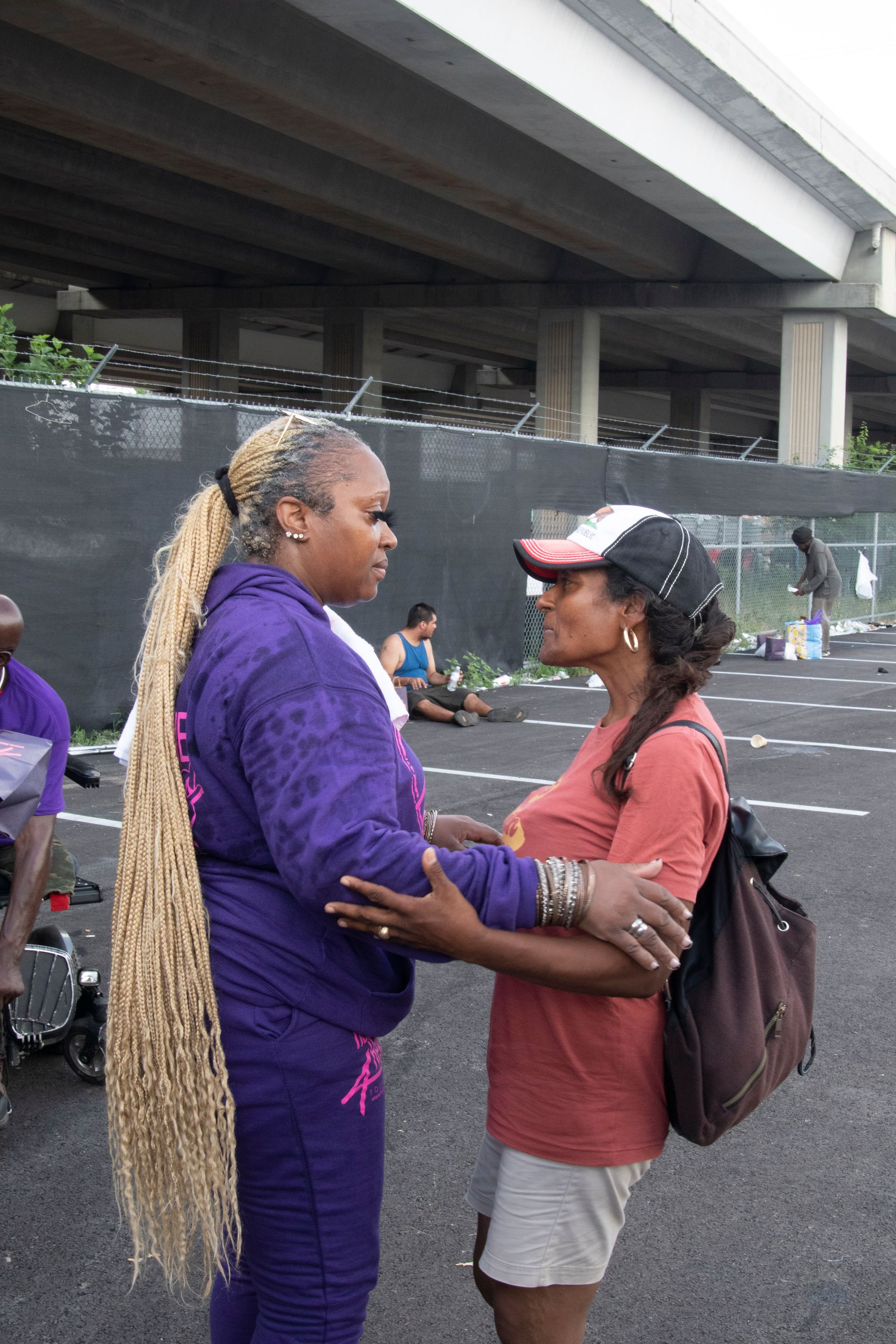 Two women are hugging each other in a parking lot under a bridge.