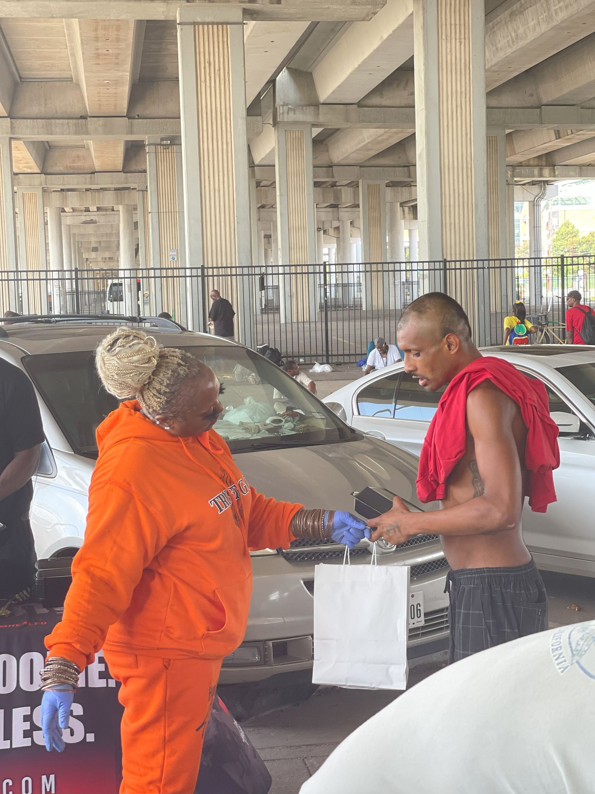 A woman in an orange hoodie is talking to a shirtless man in a parking lot.