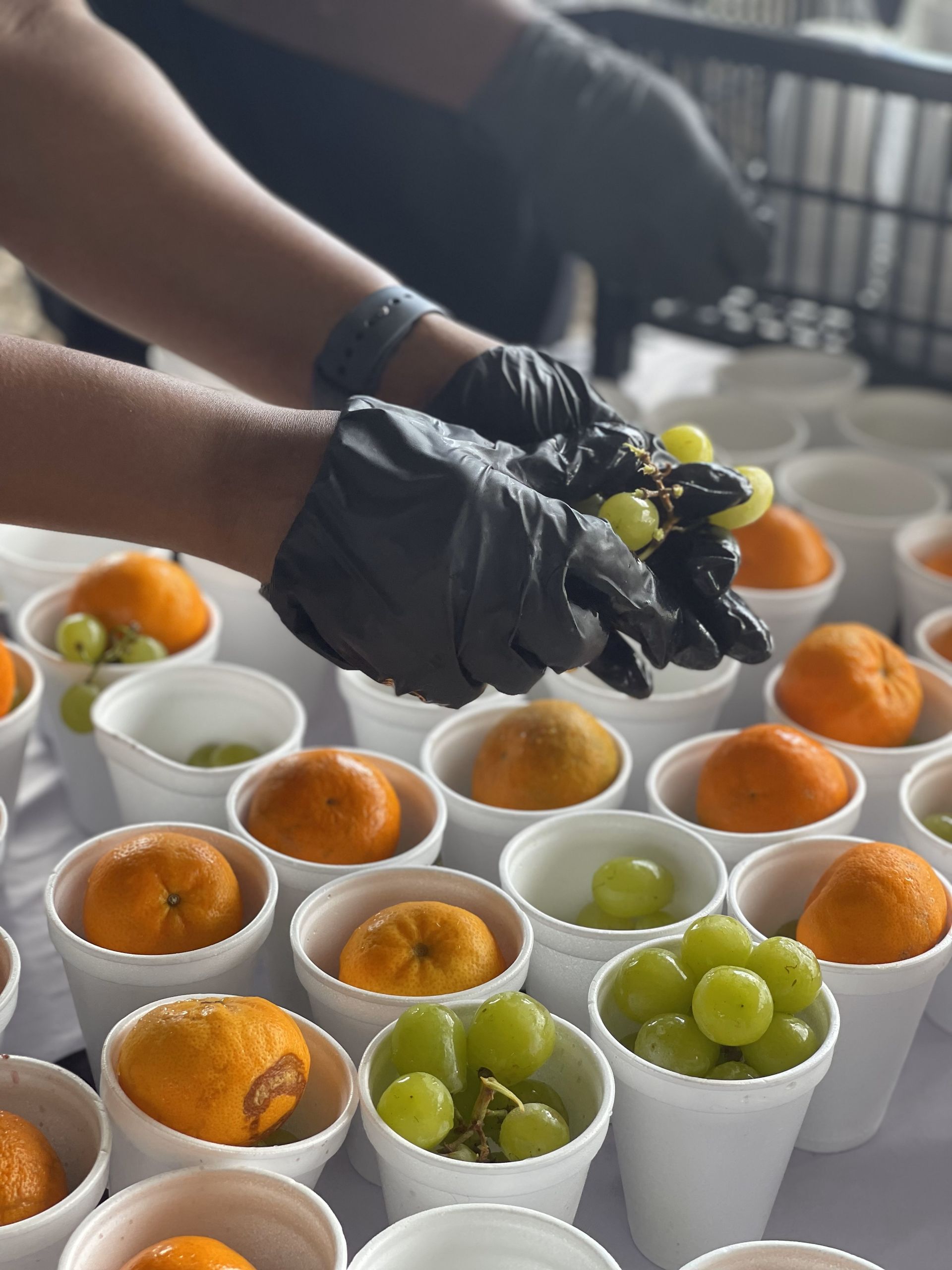 A person wearing black gloves holds a grape over a bowl of oranges