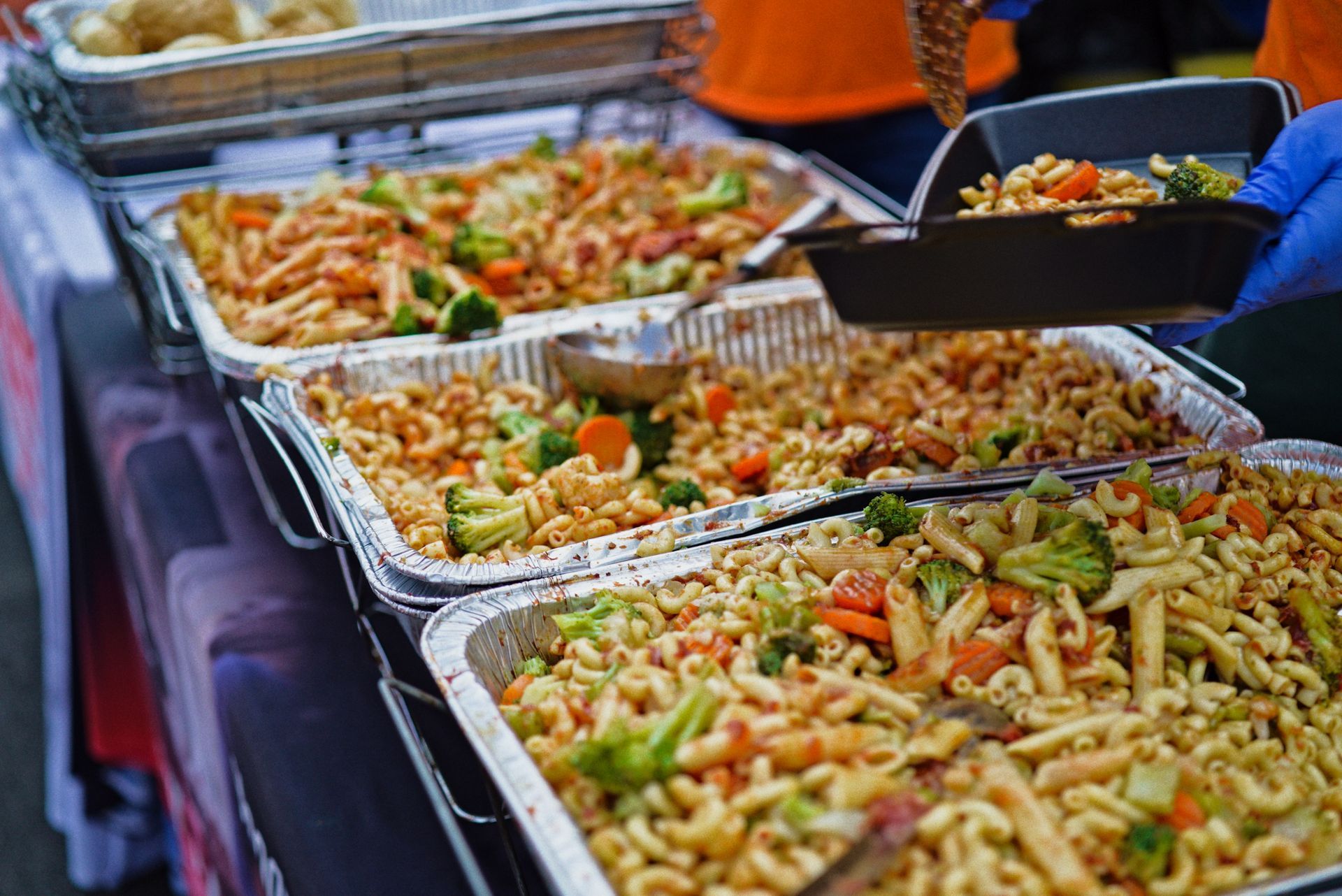 A person is taking a tray of pasta and vegetables from a buffet table.