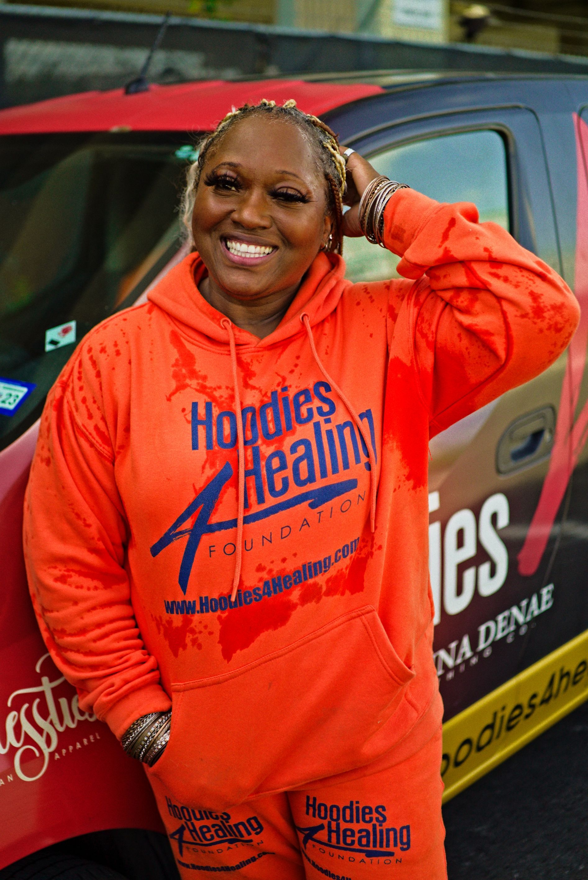 A woman in an orange hoodie is standing in front of a car.