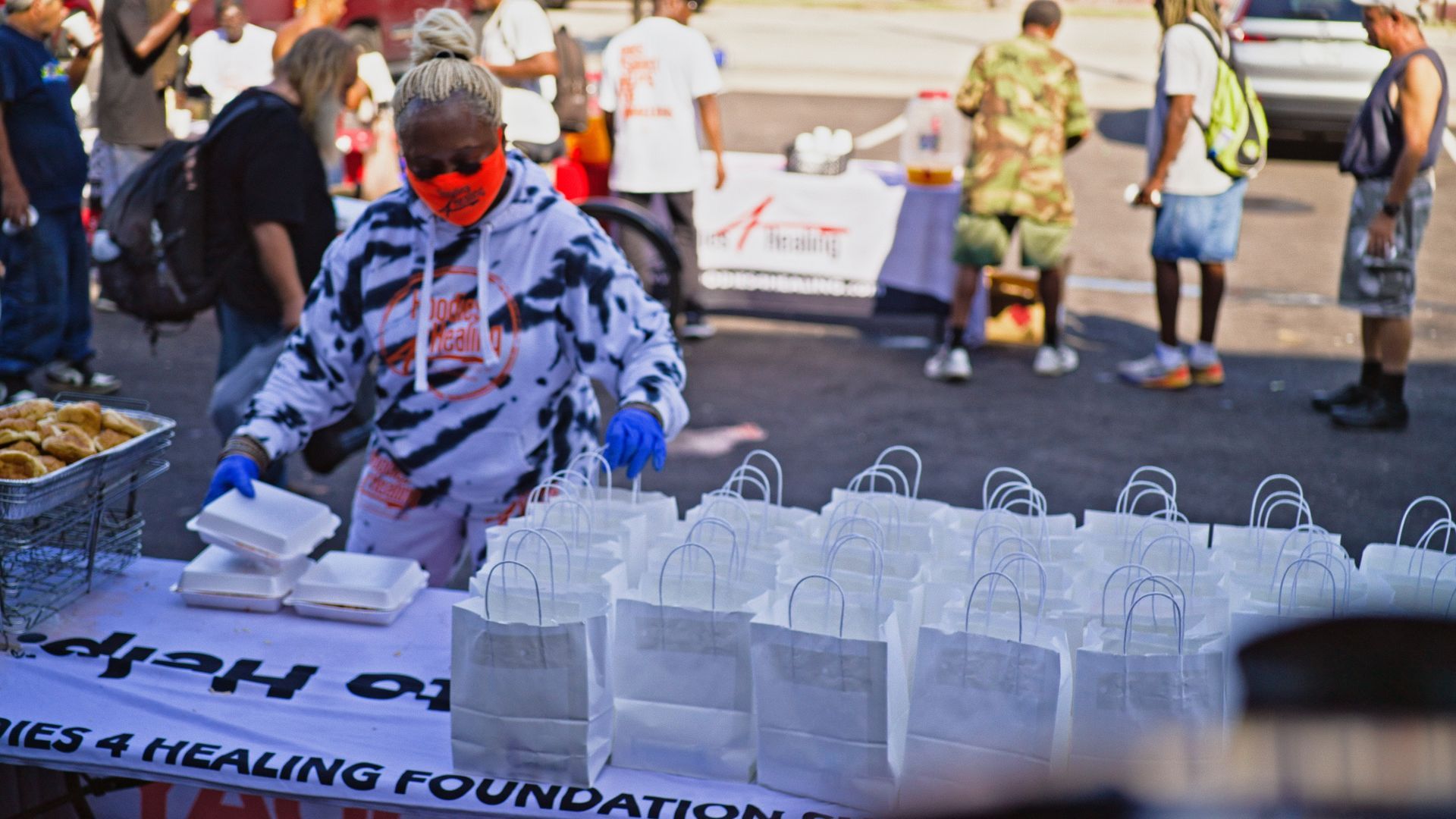 A woman wearing a mask is standing in front of a table filled with bags of food.