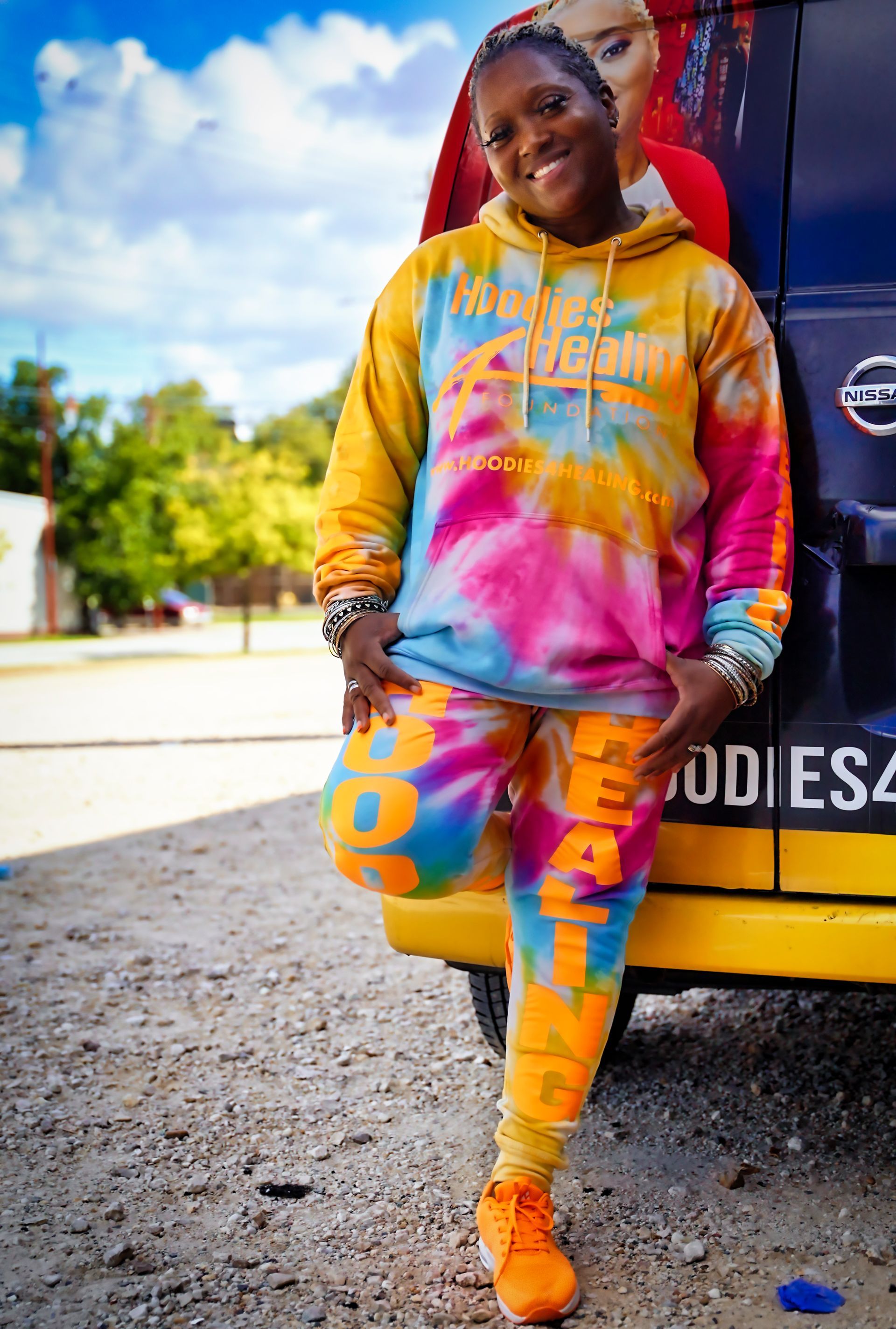 A woman in a tie dye hoodie and sweatpants is standing next to a van.