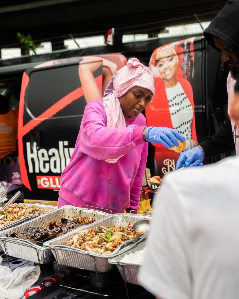 A woman in a pink sweatshirt is preparing food in front of a van that says healin ' glue