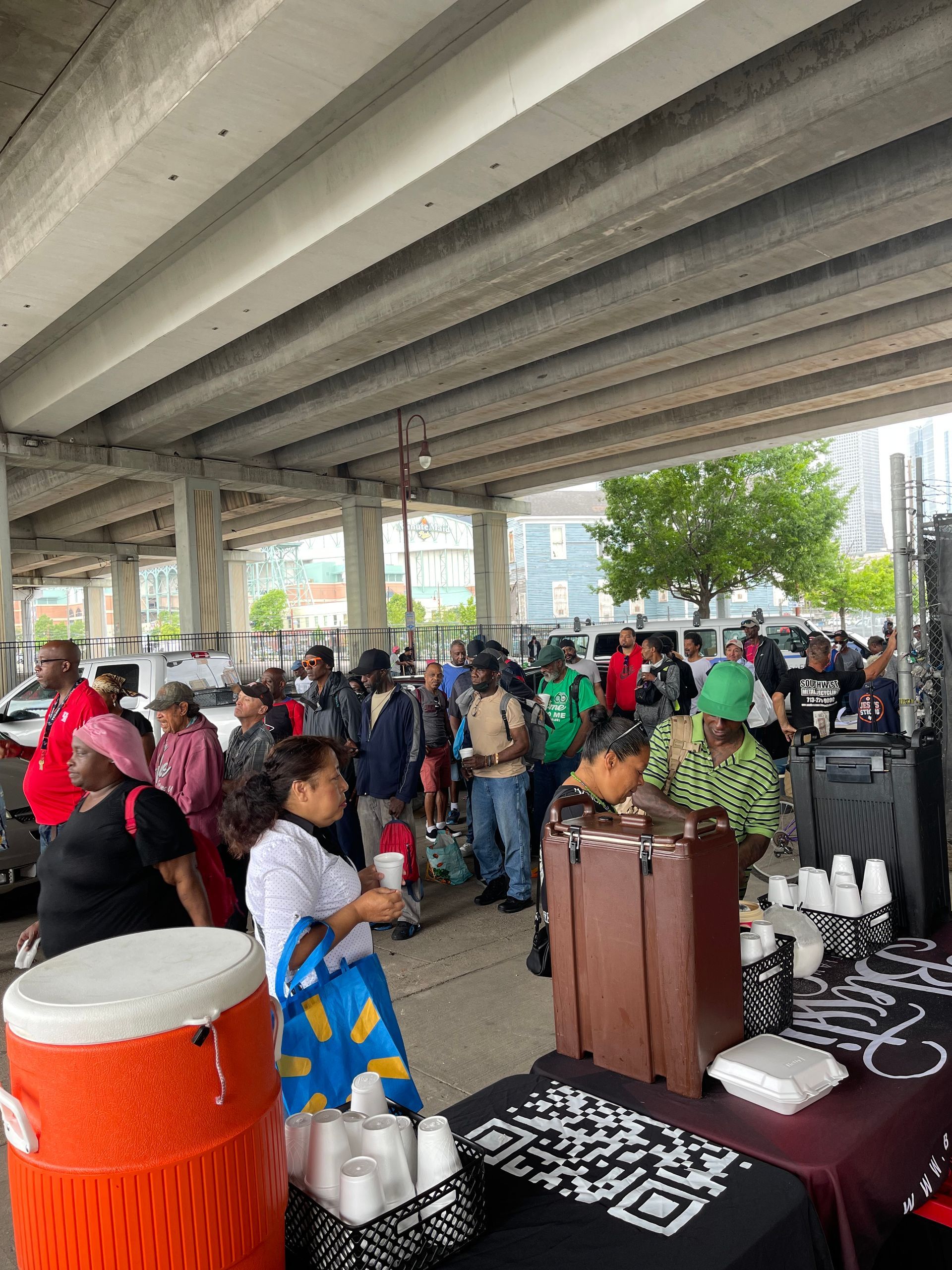 A group of people are standing around tables under a bridge.