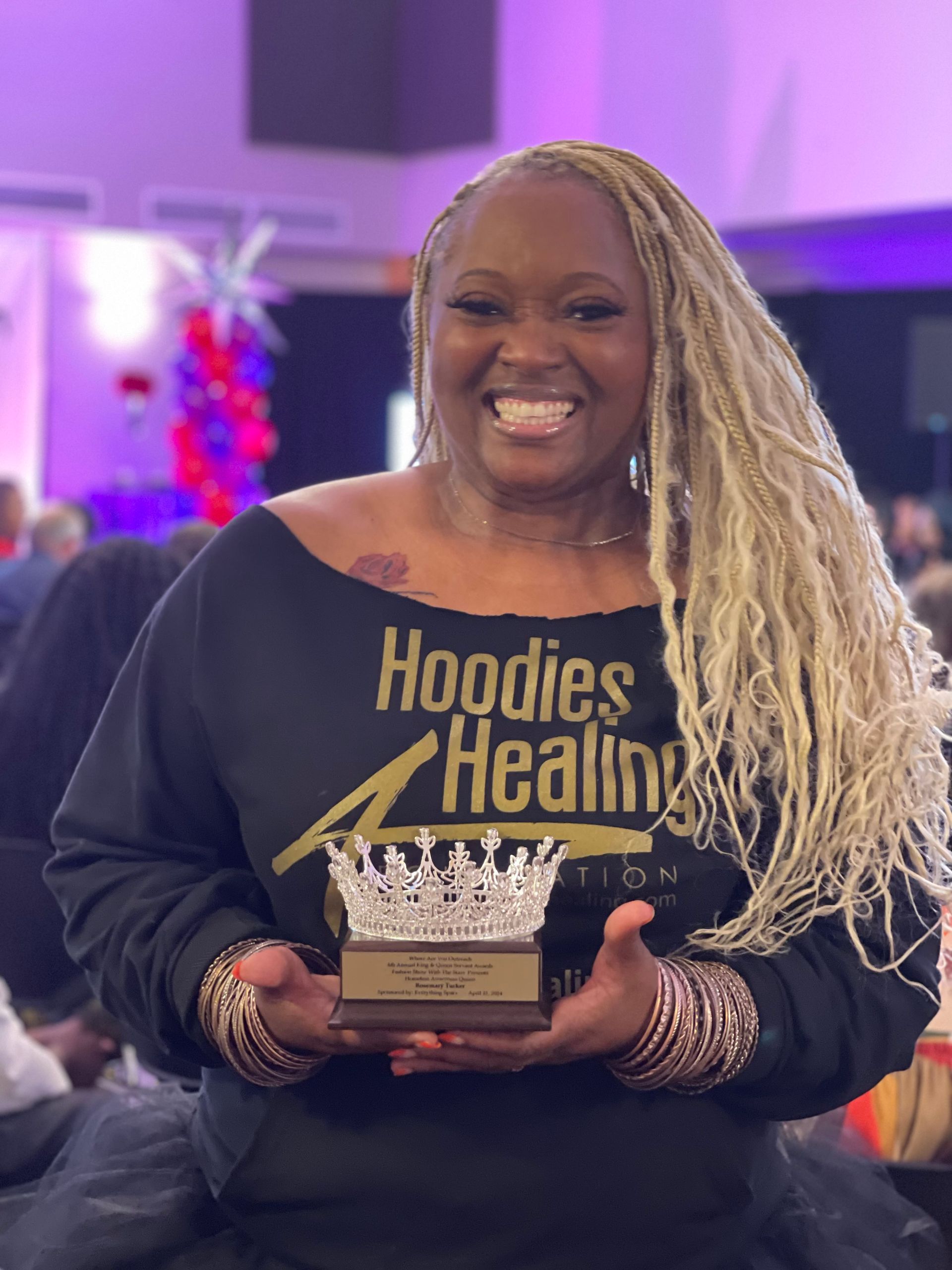 A woman wearing a black shirt that says hoodies healing is holding a trophy