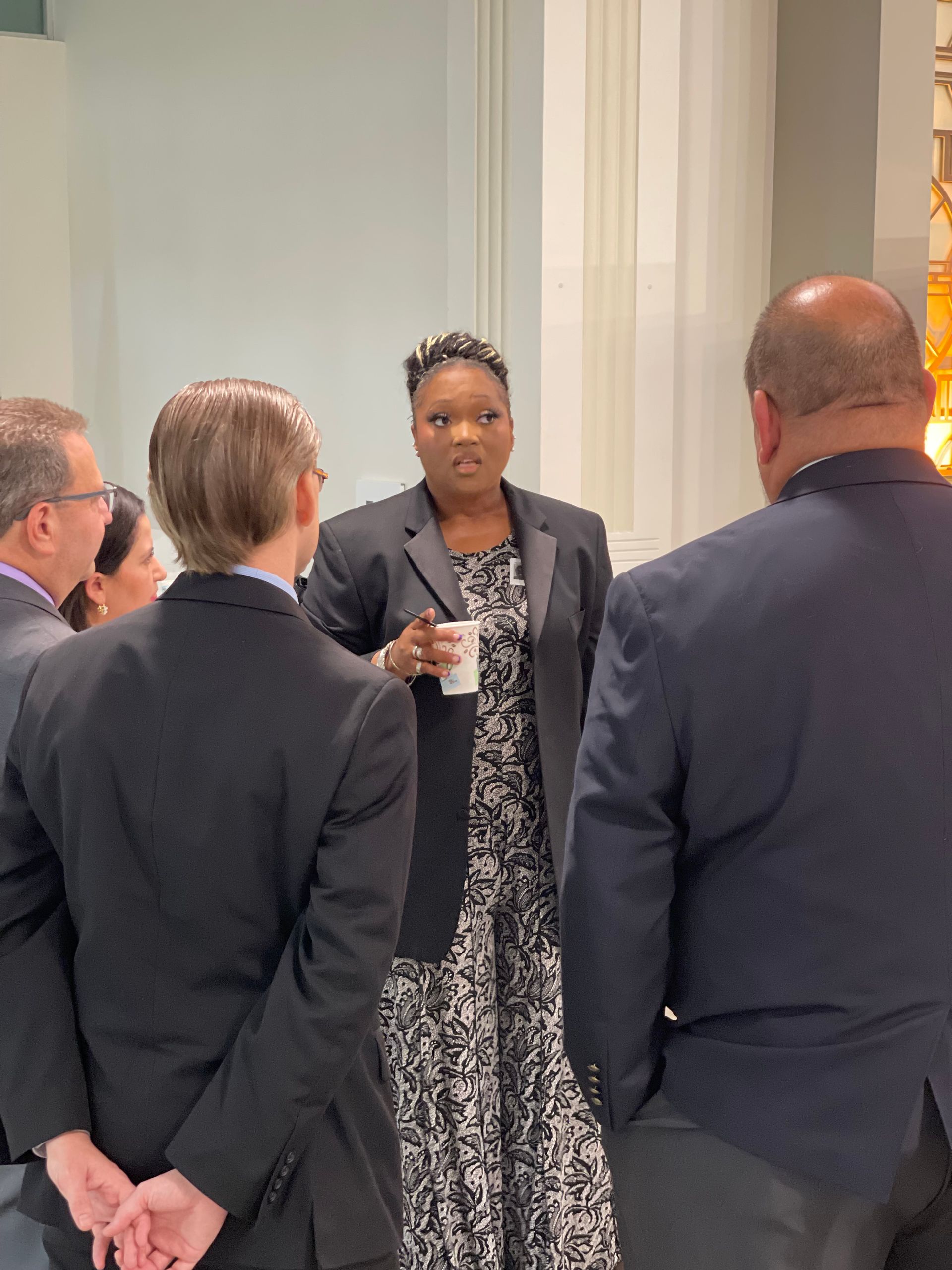 A group of people in suits are standing around a woman holding a cup of coffee.