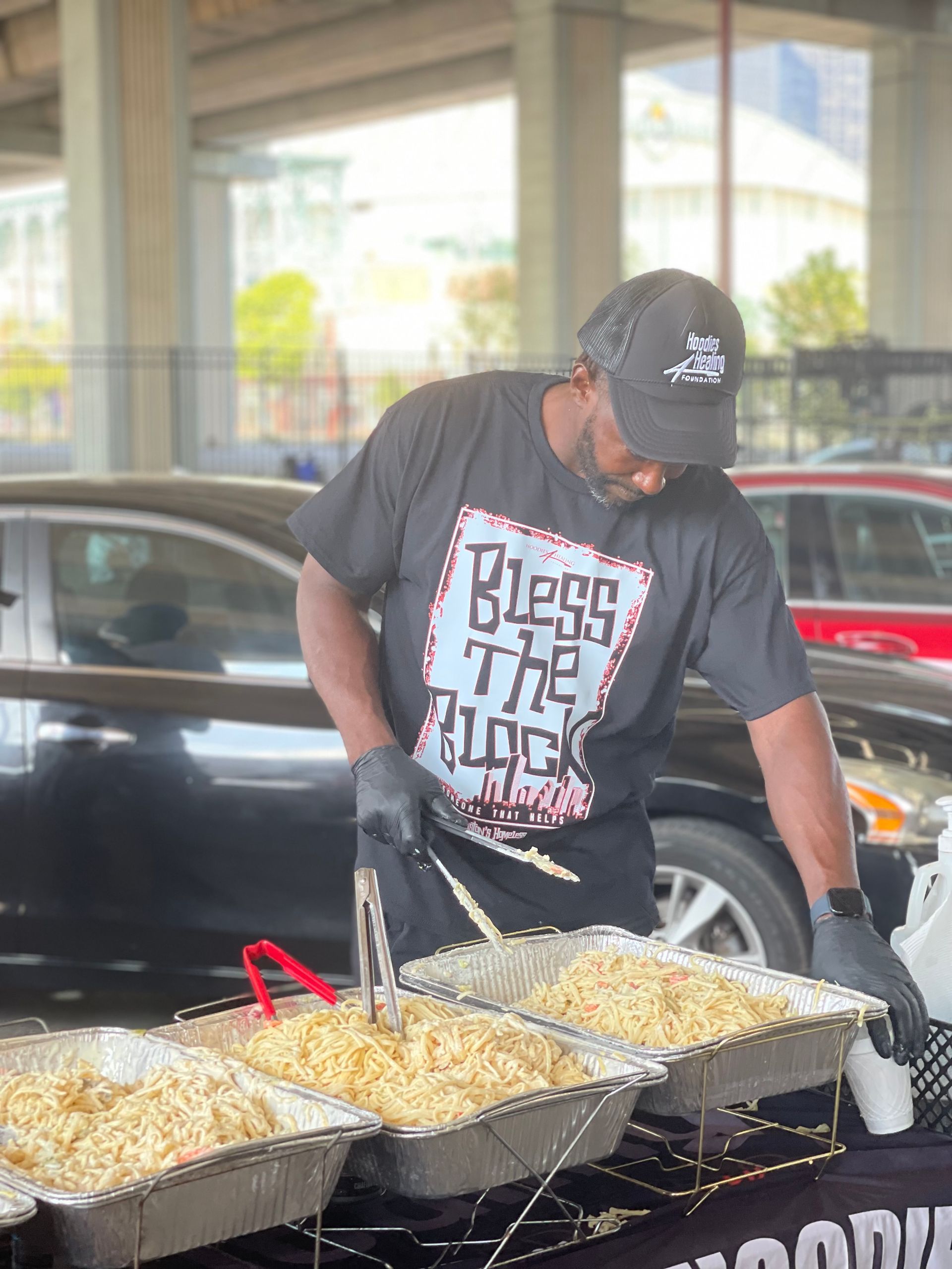 A man wearing a bless the rock shirt is cooking food in a parking lot.