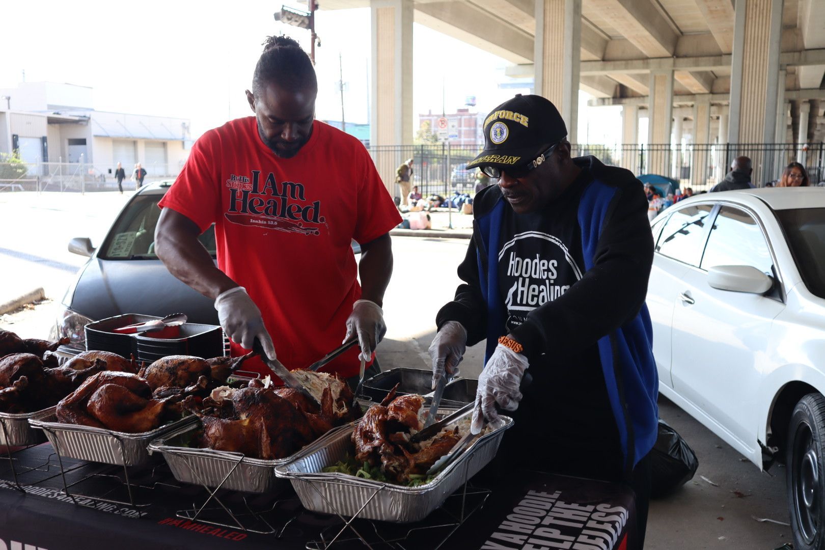Two men are preparing food in front of a white car.