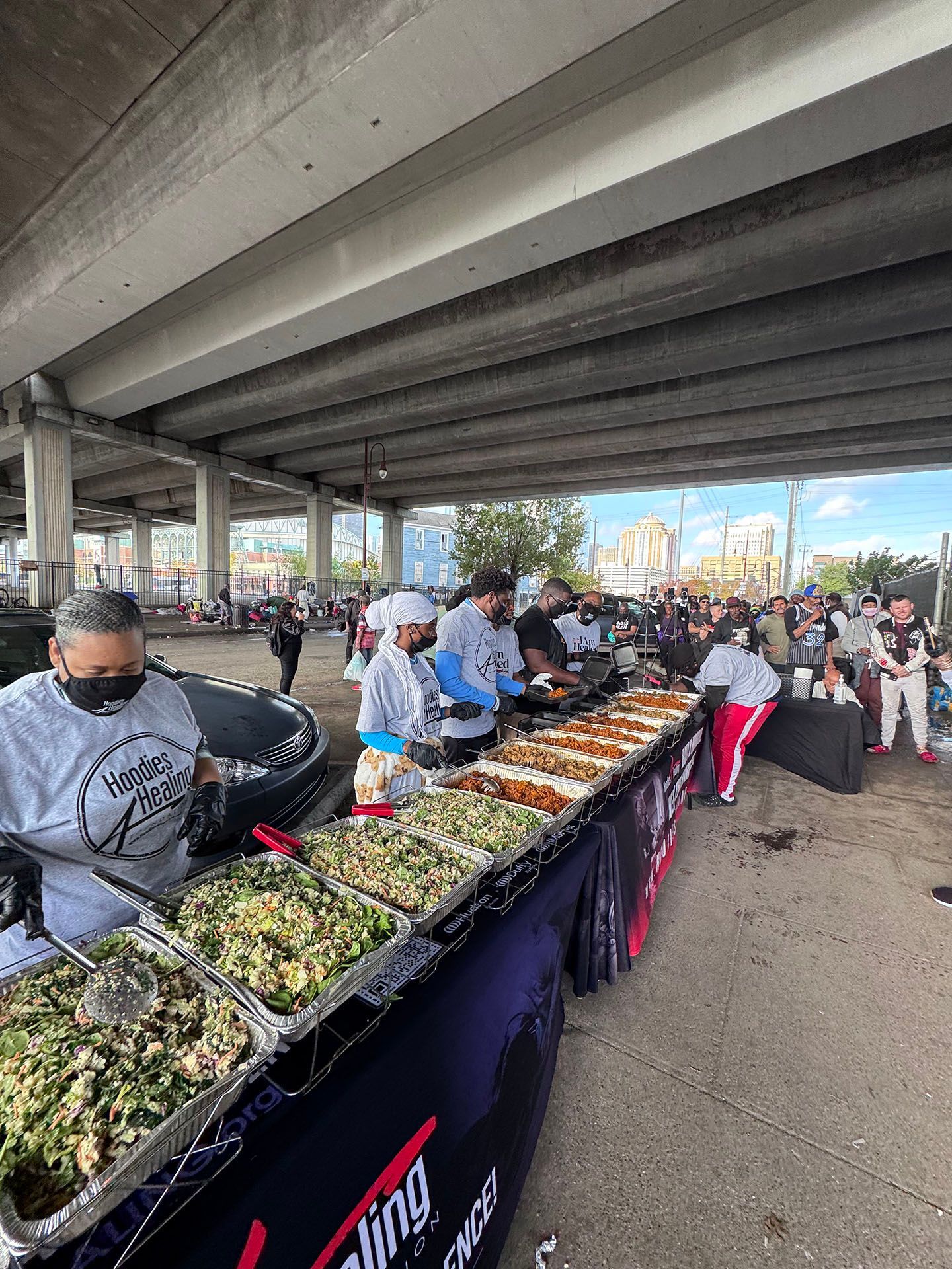 A group of people are standing around a table filled with food under a bridge.