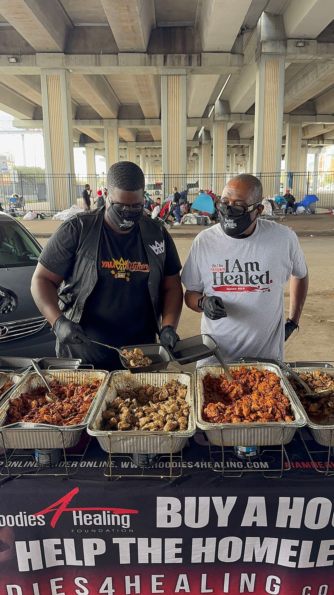 Two men are standing in front of a table filled with trays of food.