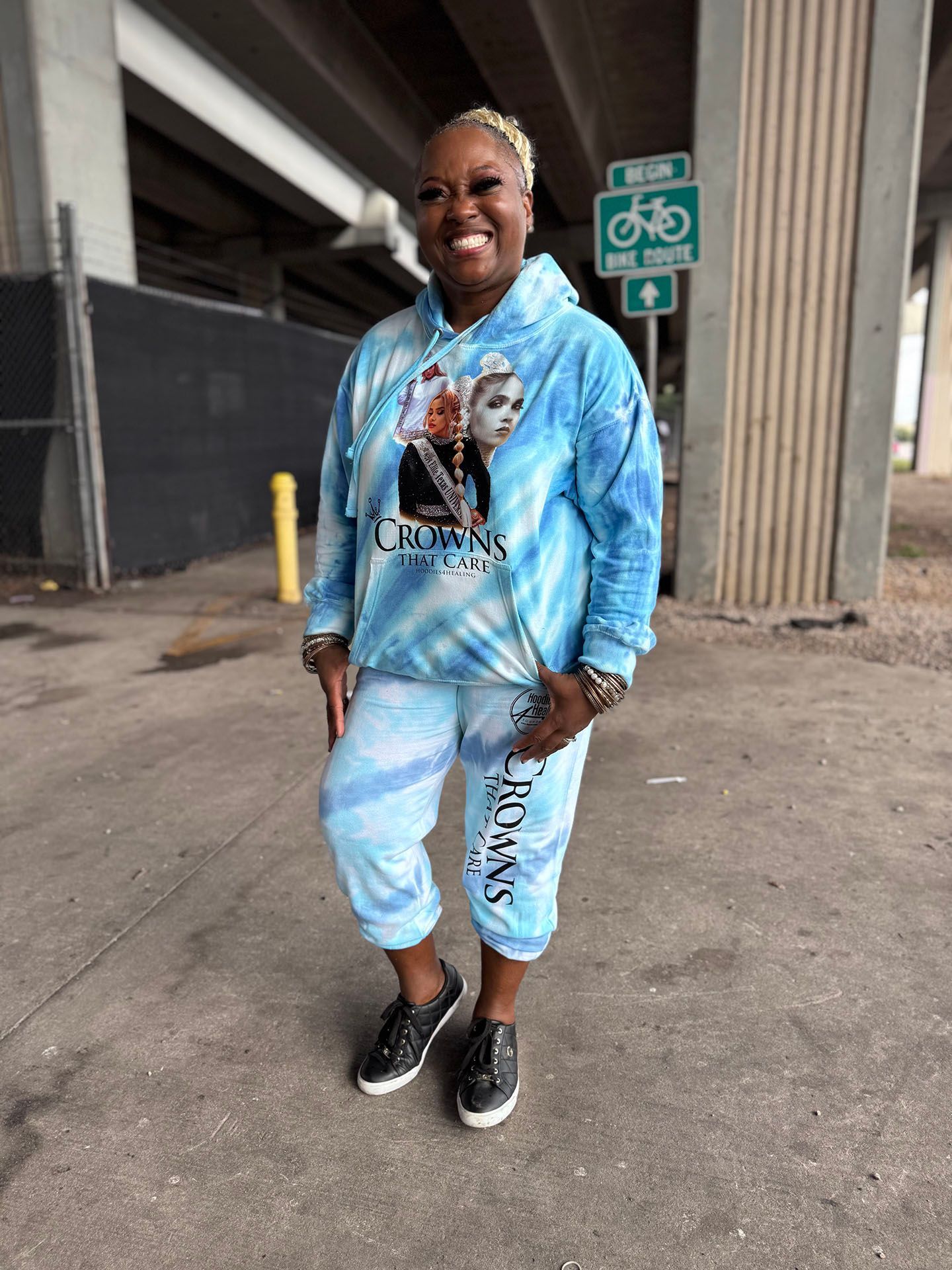 A woman wearing a tie dye hoodie and sweatpants is standing in front of a bridge.