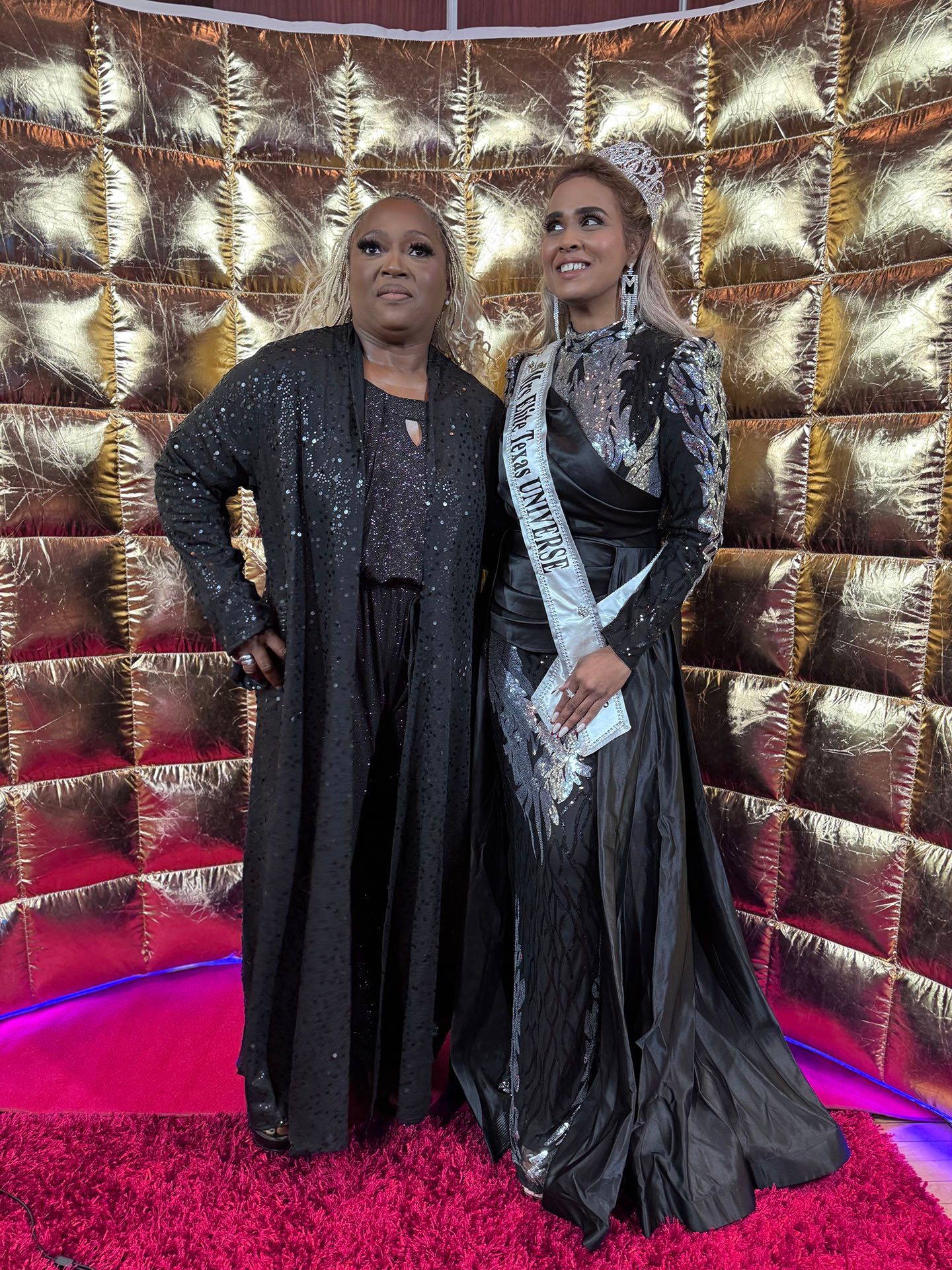 Two women in black dresses are standing next to each other in front of a gold wall.