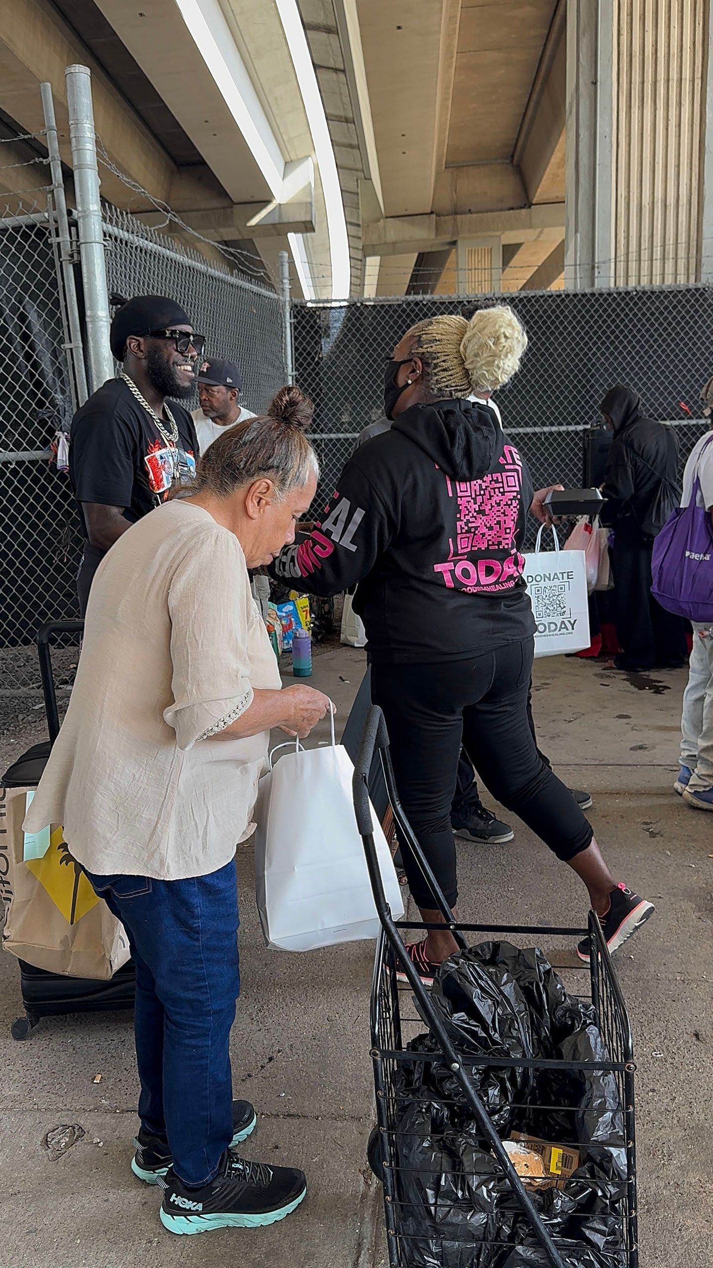 A group of people are standing in a parking lot under a bridge.