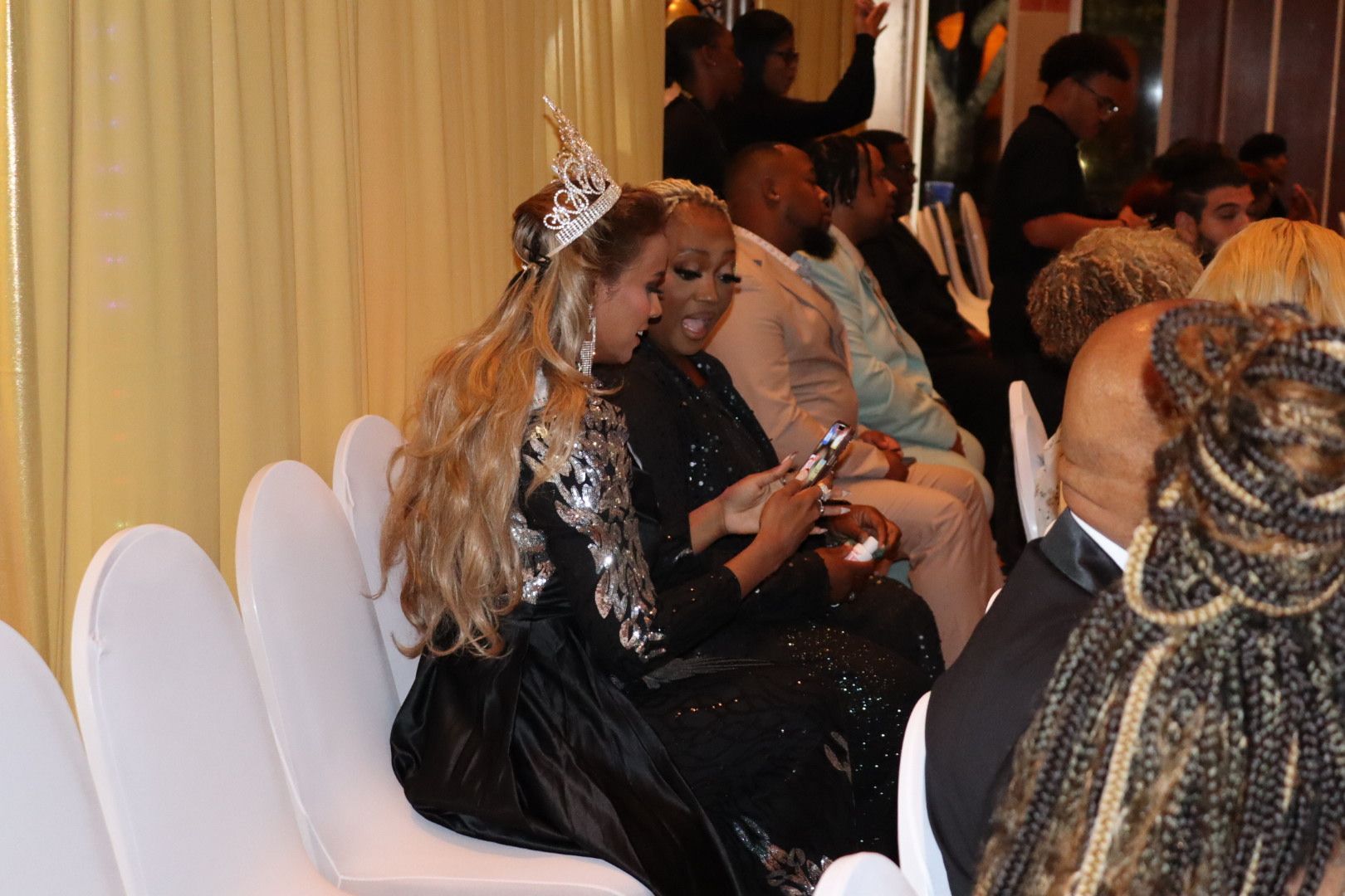 A woman wearing a tiara is sitting in a row of chairs with other people