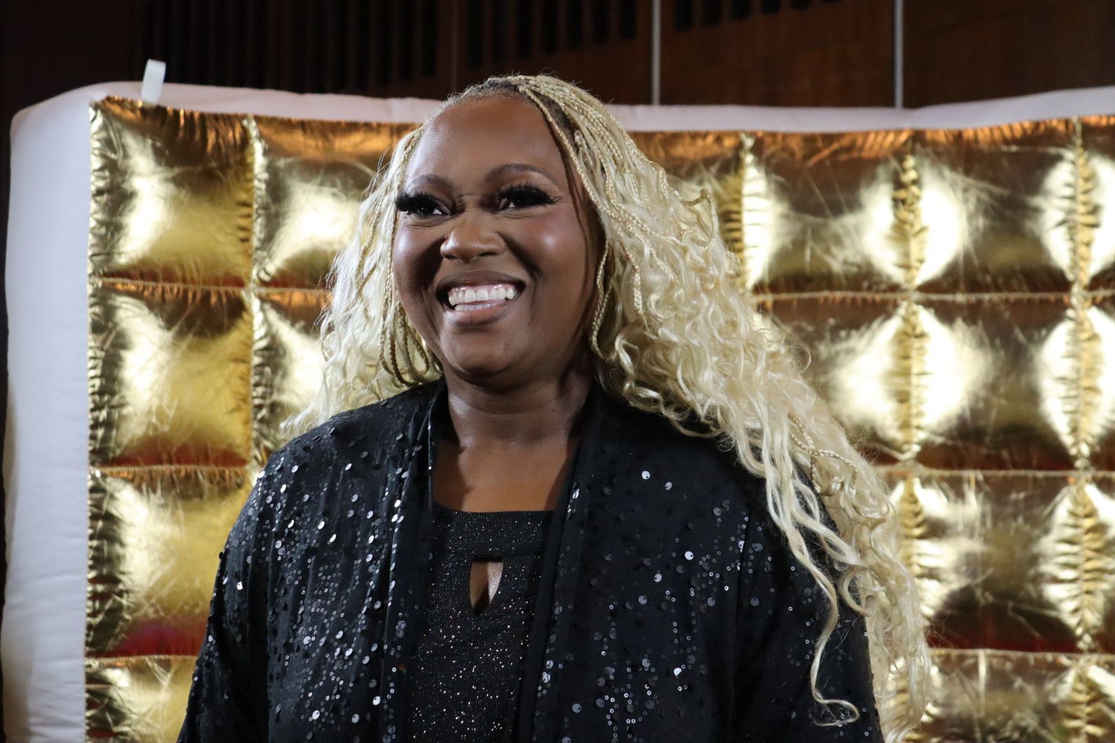 A woman with blonde hair is smiling in front of a gold wall
