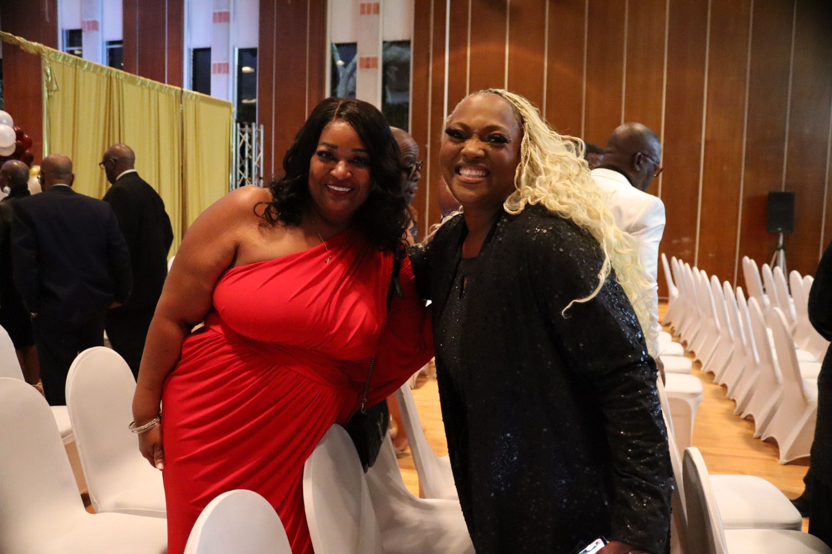Two women are posing for a picture in a room with white chairs.