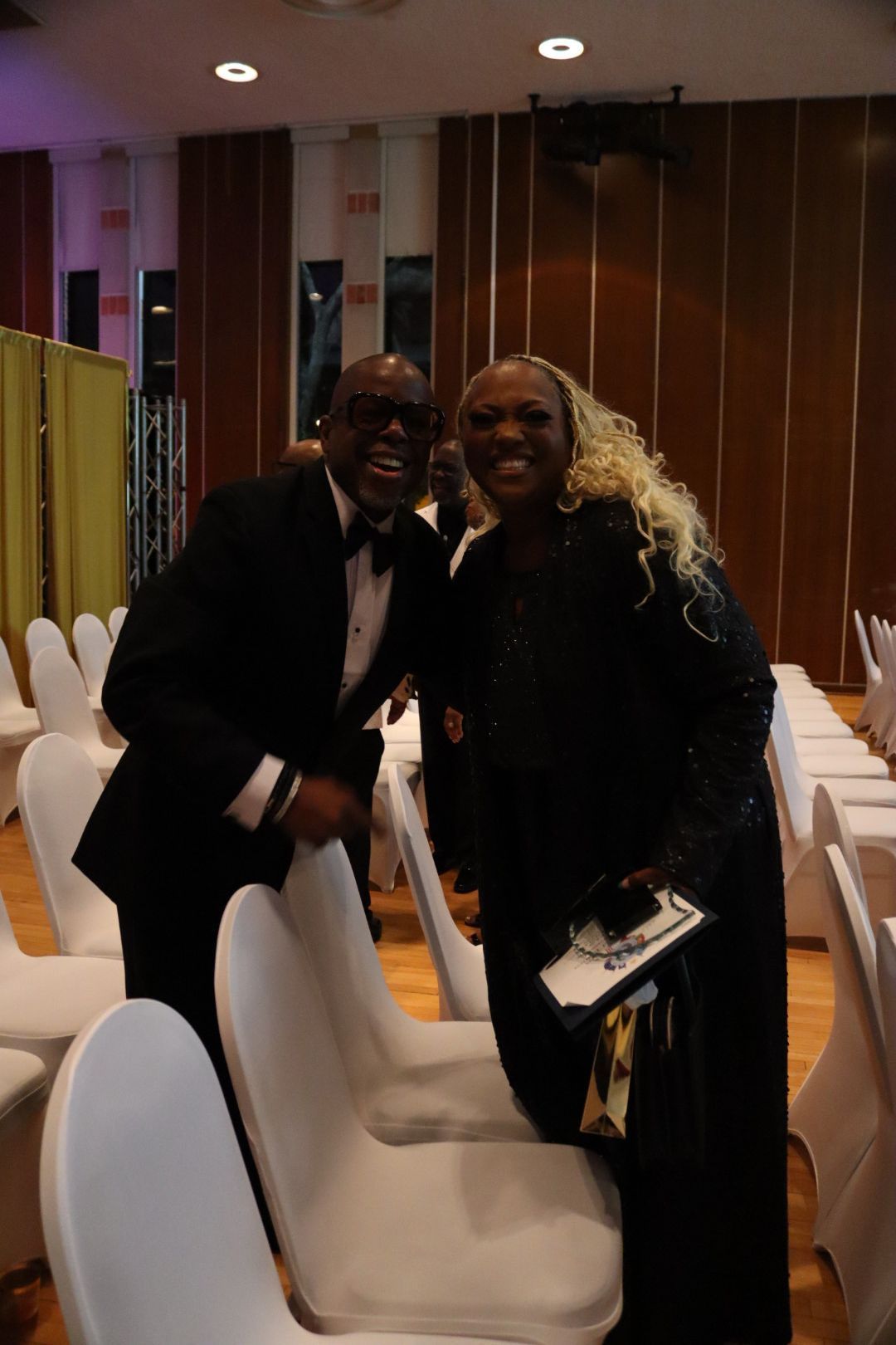 A man and a woman are posing for a picture in a room with white chairs.