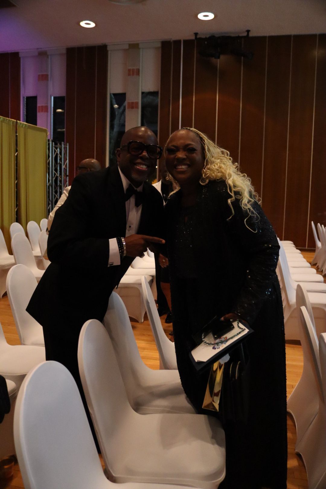 A man and a woman are posing for a picture in a room with white chairs.