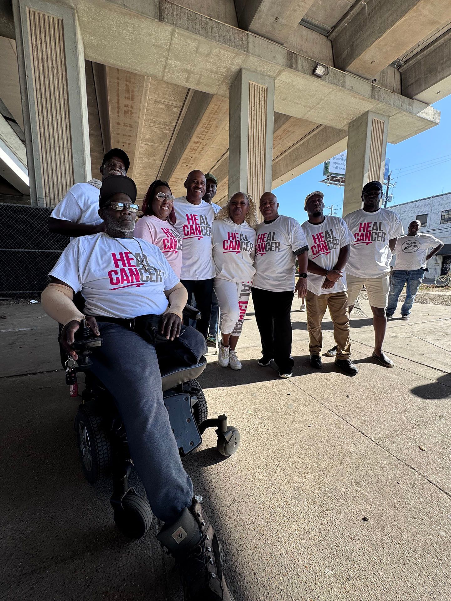 A man in a wheelchair is sitting in front of a group of people standing under a bridge.
