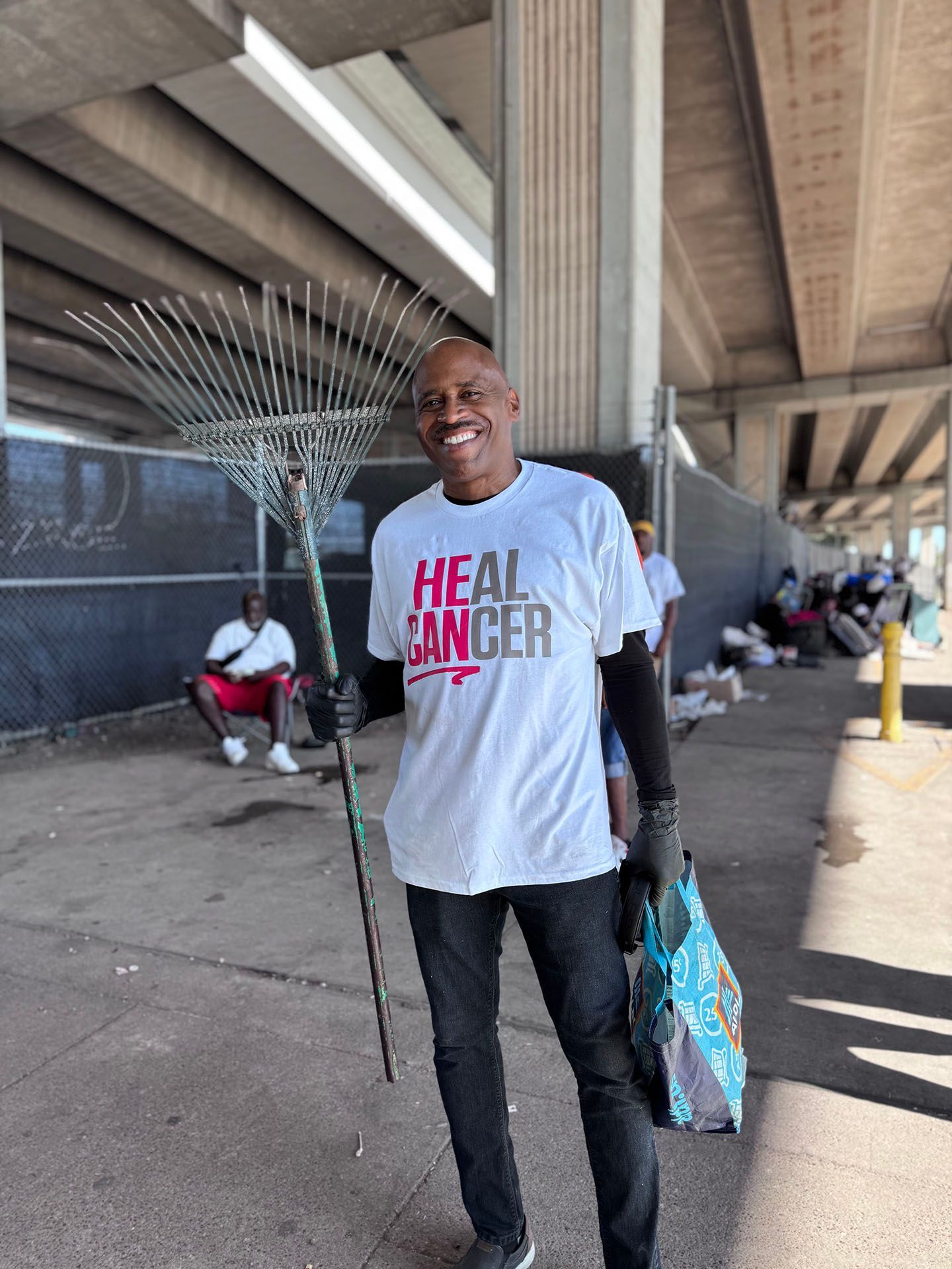 A man wearing a white t-shirt that says `` heal cancer '' is holding a rake and a bag.