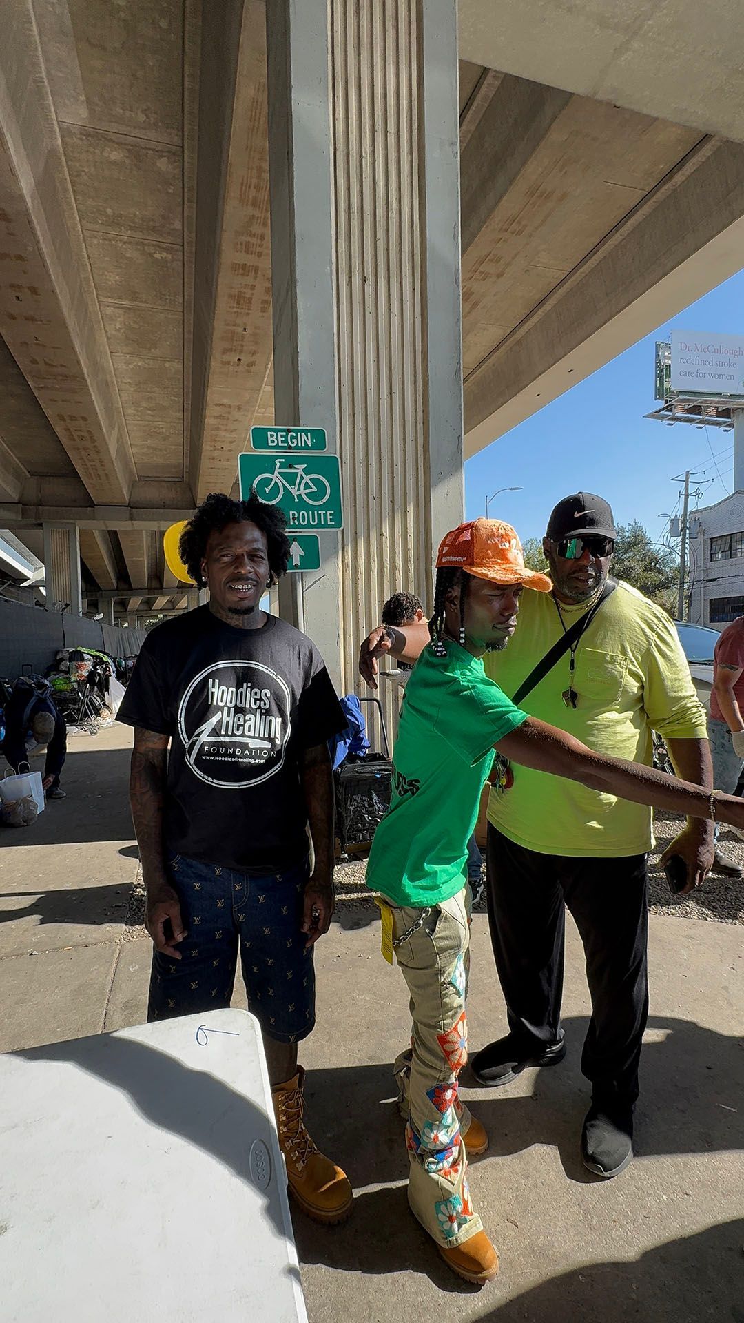 A group of people are standing on a sidewalk under a bridge.