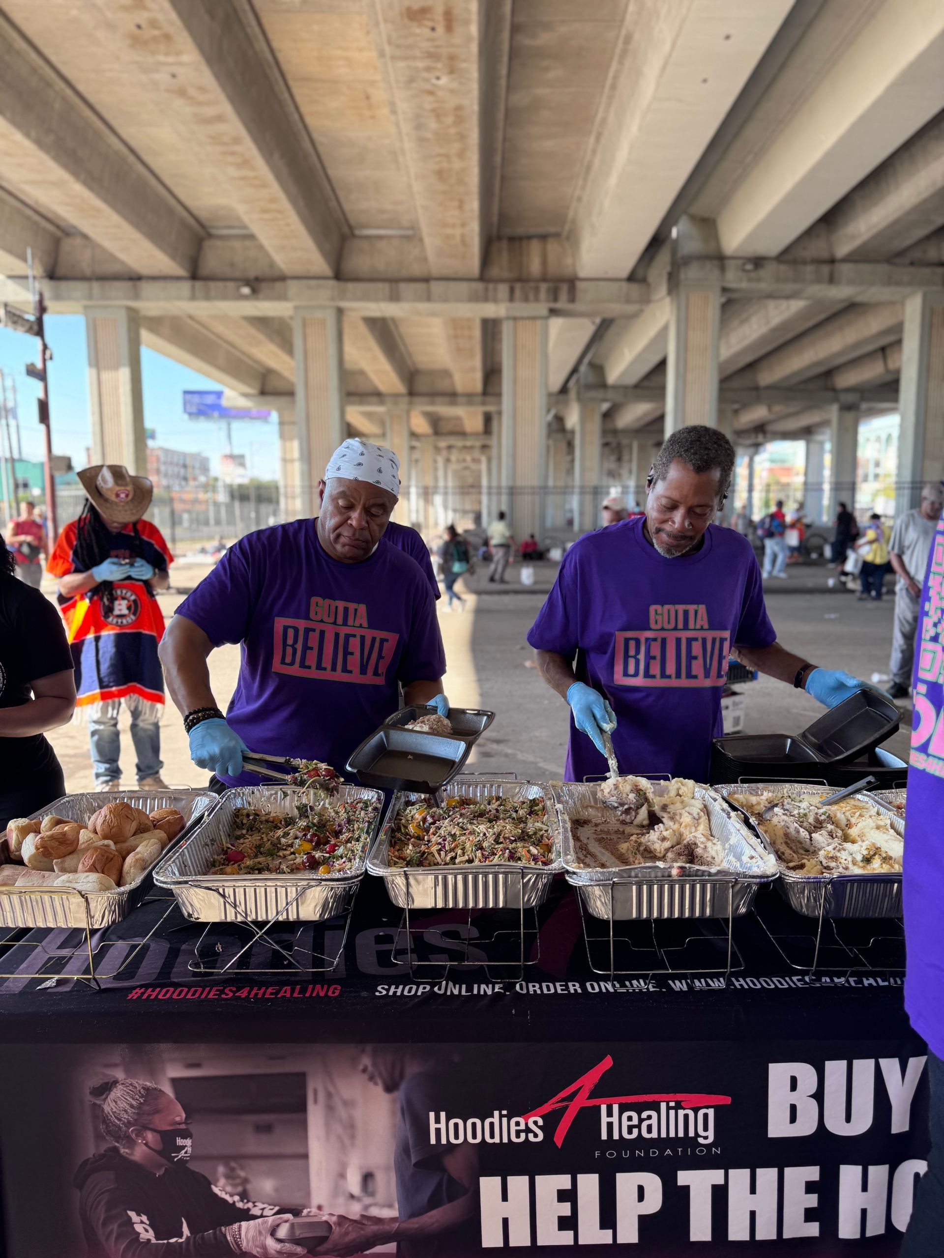 Two men in purple shirts are preparing food at a table.
