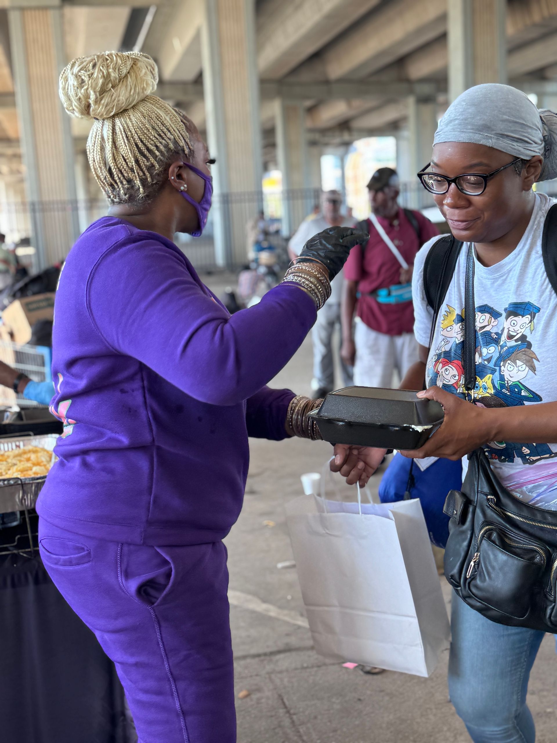 A woman in a purple outfit is giving a box of food to another woman.
