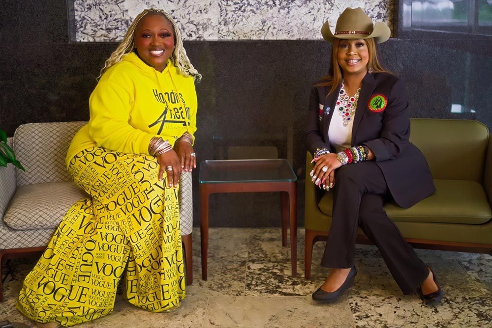 Two women are sitting next to each other on a couch . one of the women is wearing a cowboy hat.