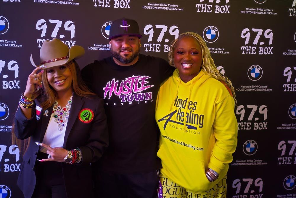 Three people are posing for a picture in front of a wall that says 97.9 the box.