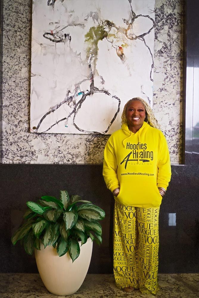A woman in a yellow hoodie is standing next to a potted plant.