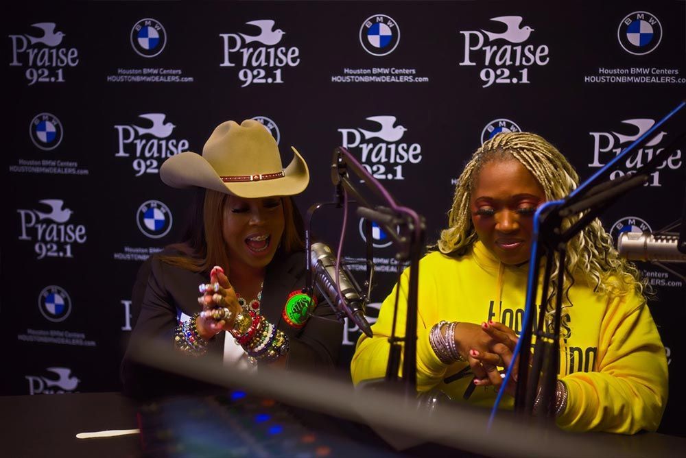 Two women are sitting in front of microphones in front of a wall that says praise 92.1.