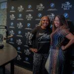 Two women are standing next to each other in front of a wall.