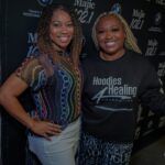 Two women are posing for a picture together in front of a black wall.
