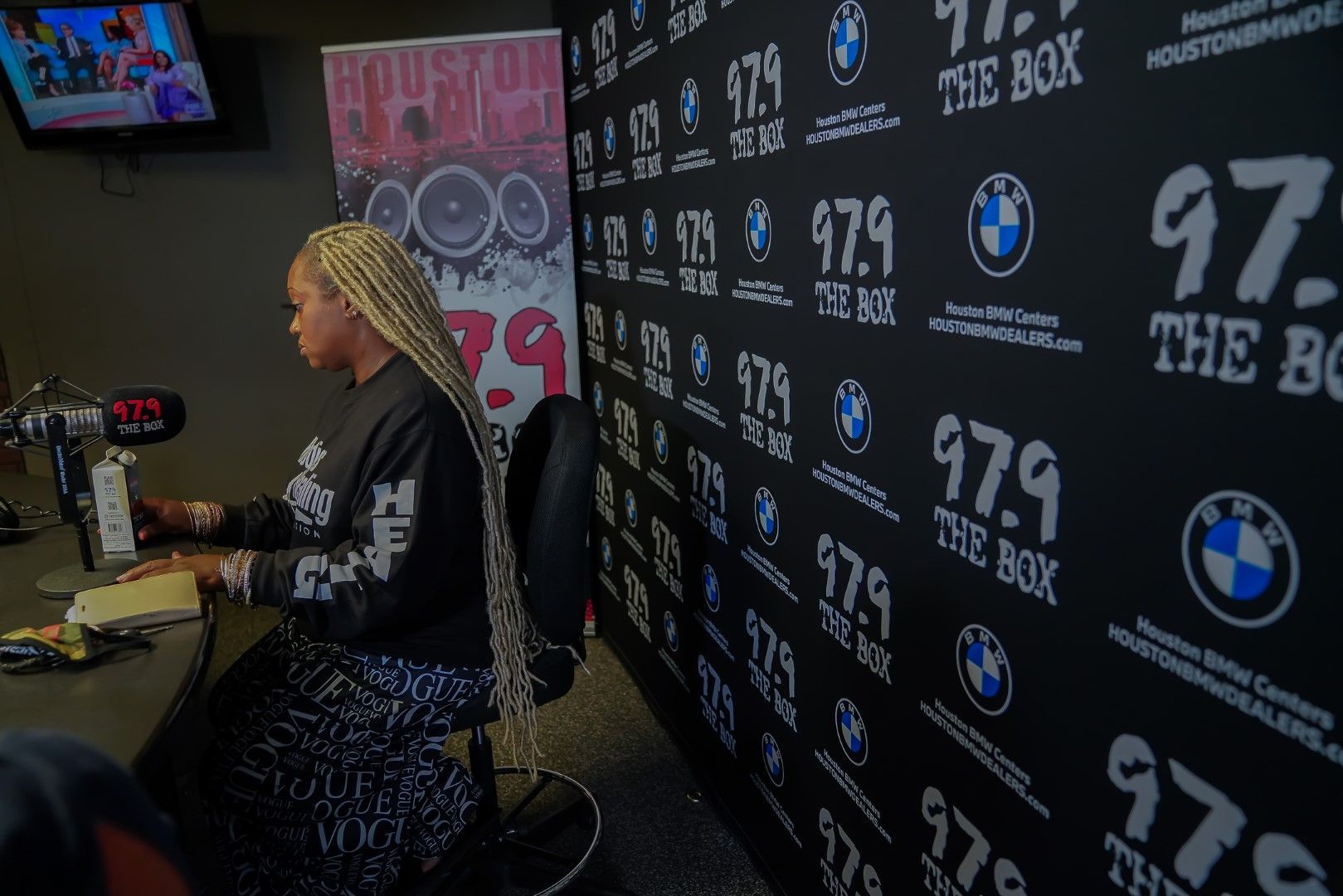 A woman is sitting at a desk in front of a wall that says 97.9 the box.