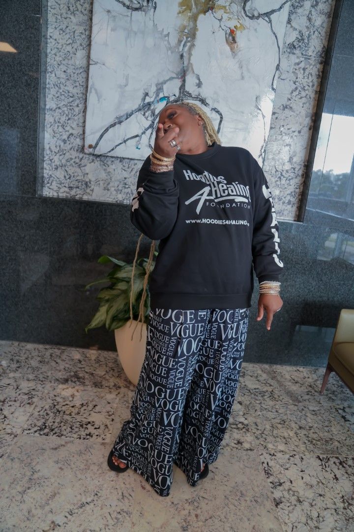 A woman wearing a black sweatshirt and wide leg pants is standing in front of a painting.