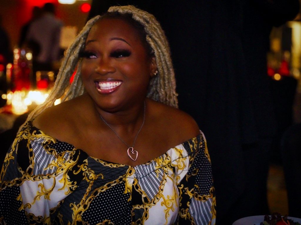 A woman with blonde braids is smiling and wearing a black and gold dress.