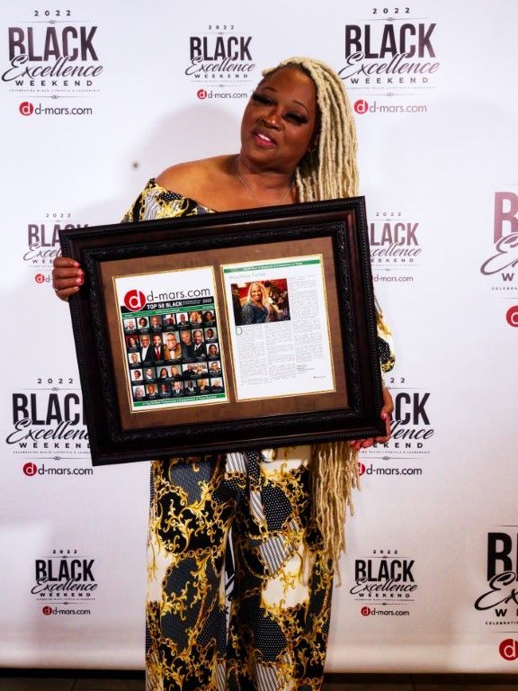 A woman is holding a framed picture in front of a wall that says black excellence