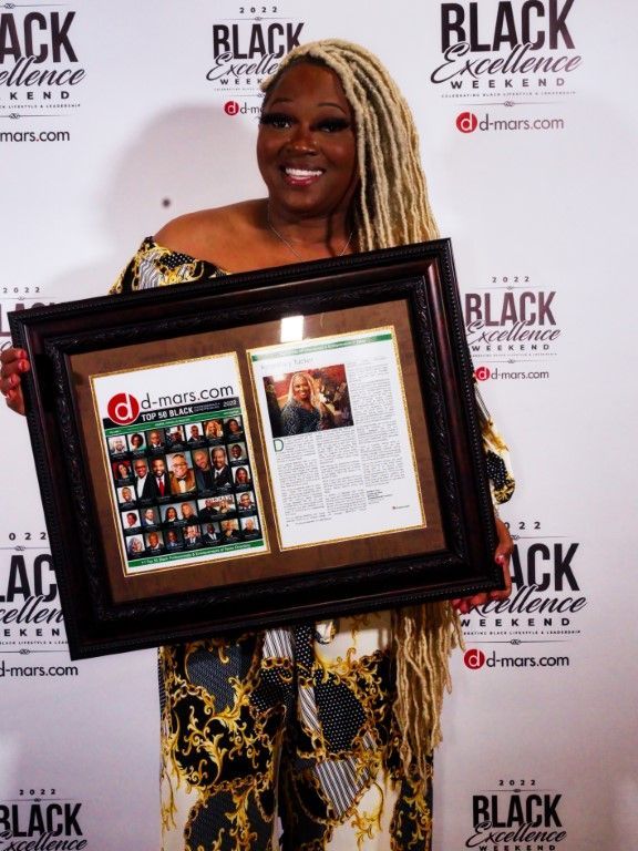 A woman is holding a framed picture in front of a wall that says black excellence