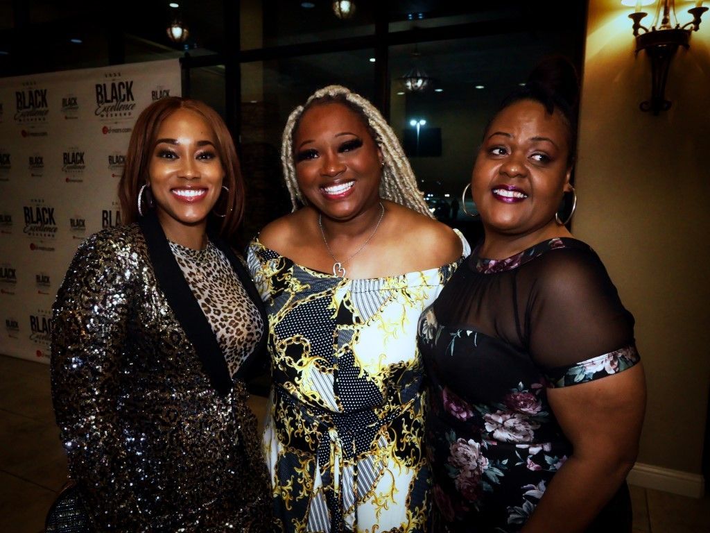 Three women are posing for a picture in front of a wall that says black