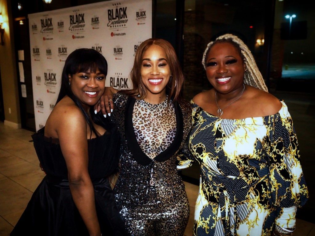 Three women are posing for a picture in front of a wall that says black