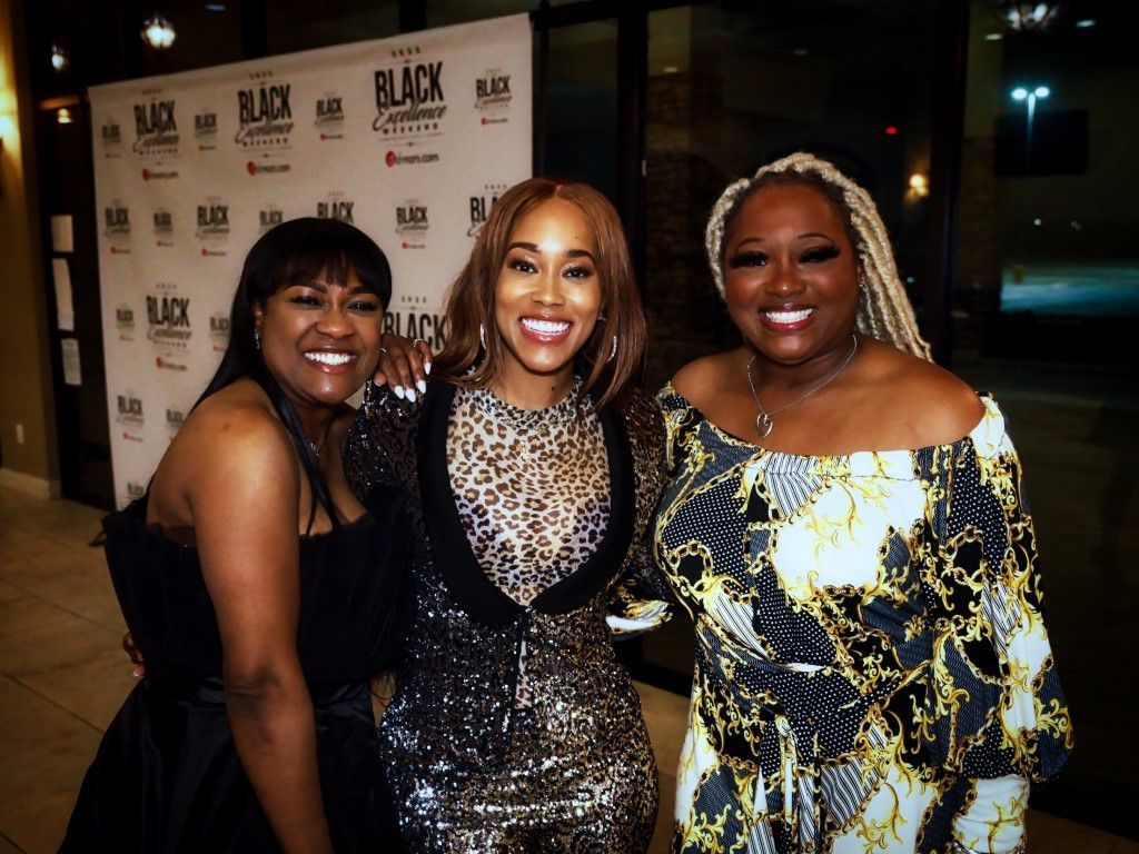 Three women are posing for a picture in front of a wall that says black